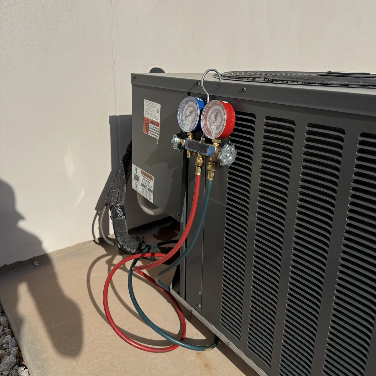 A manifold gauge set hanging from the service valves of an outdoor air conditioning condenser beside a stucco wall