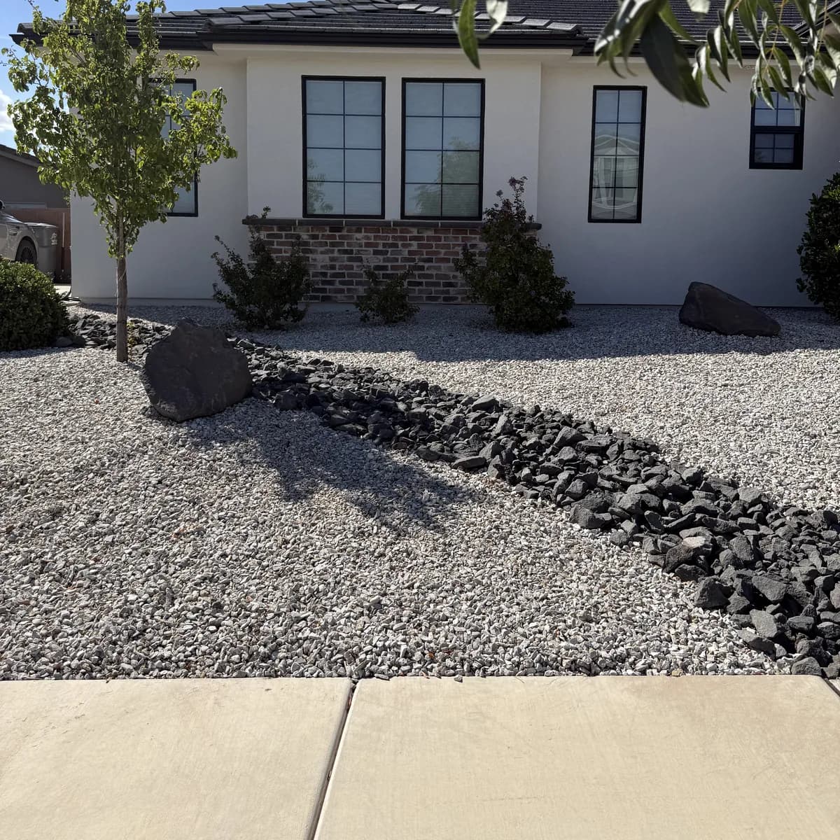 A front yard in St. George converted to pale gravel with a dry creek of dark rock, young trees and low shrubs along a white stucco house
