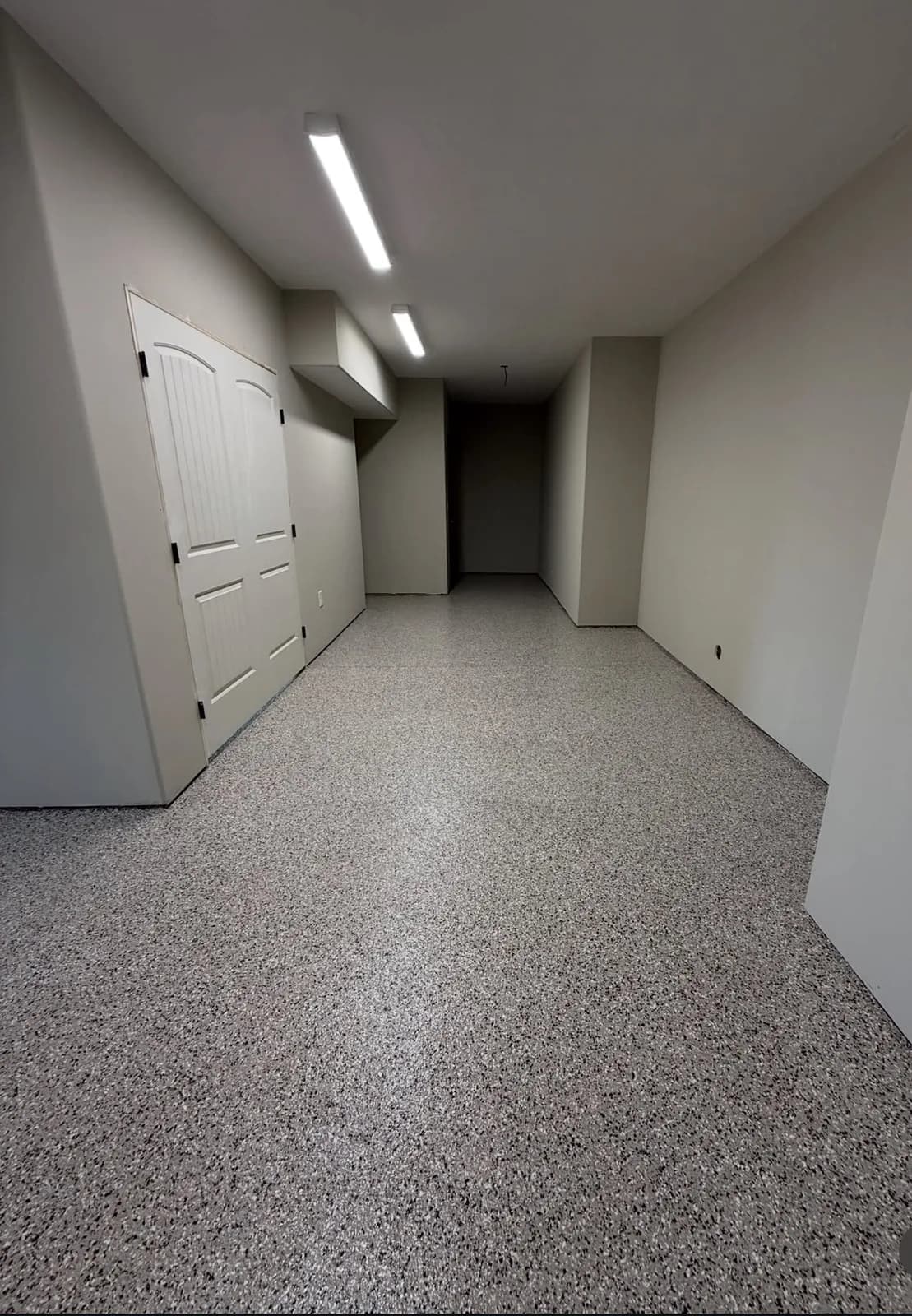 A basement corridor finished in a fine tan and brown flake floor, running past white-painted walls to an open utility room at the end