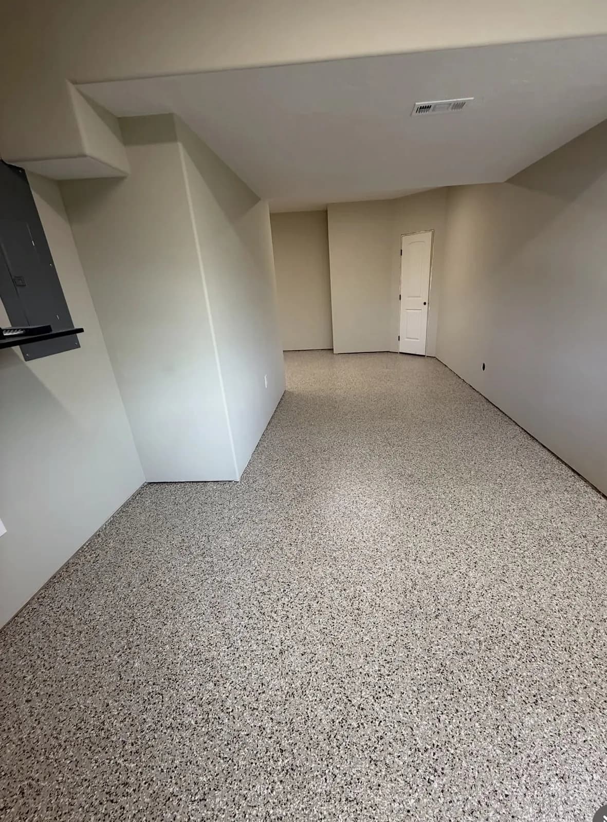 A basement room with a fine pale tan and gray flake floor throughout, white walls, an electrical panel on the left and a white door at the far end
