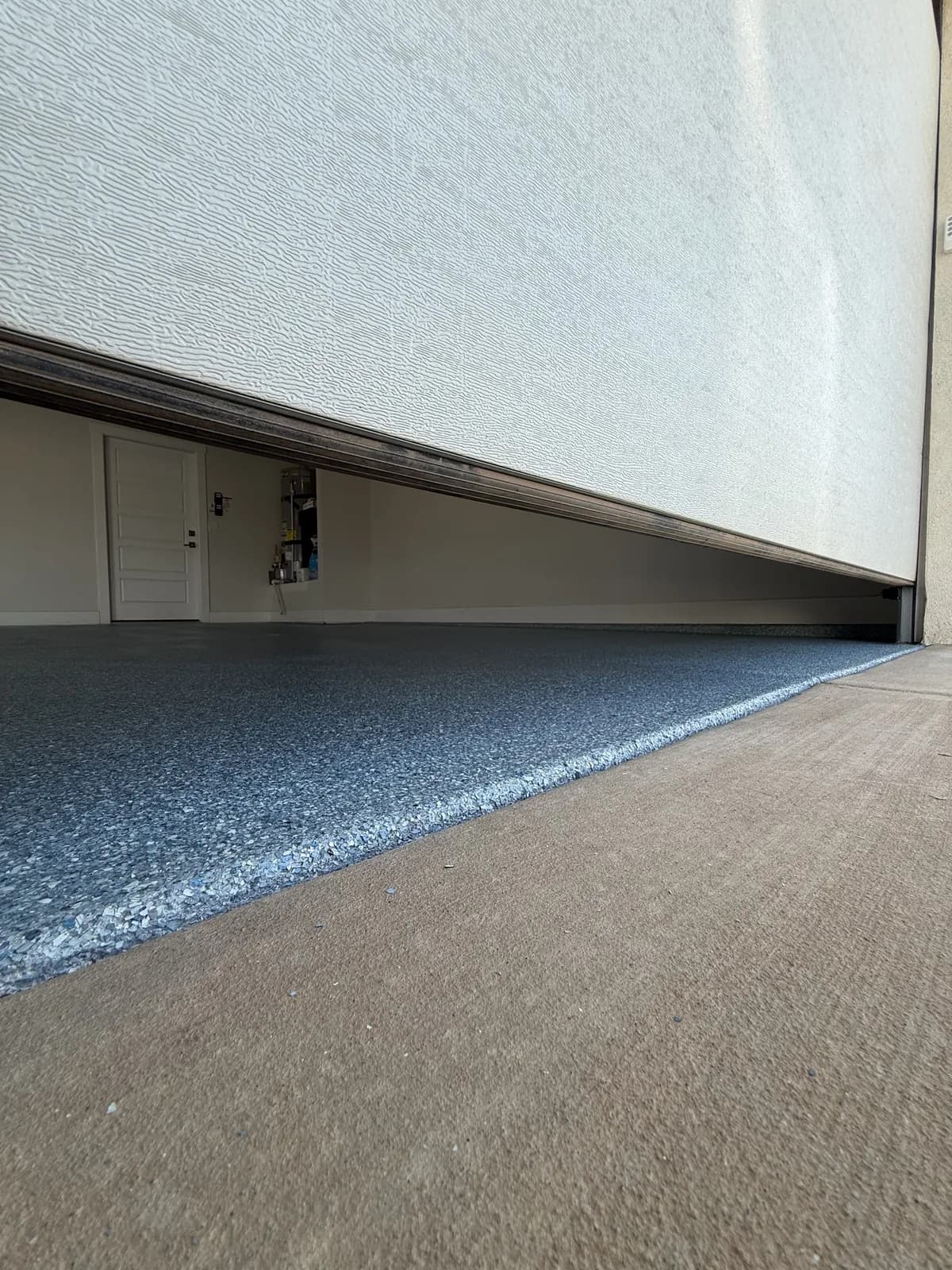 A ground-level view along the threshold of a garage door, showing a blue and white flake floor inside ending in a clean edge against the plain concrete driveway outside