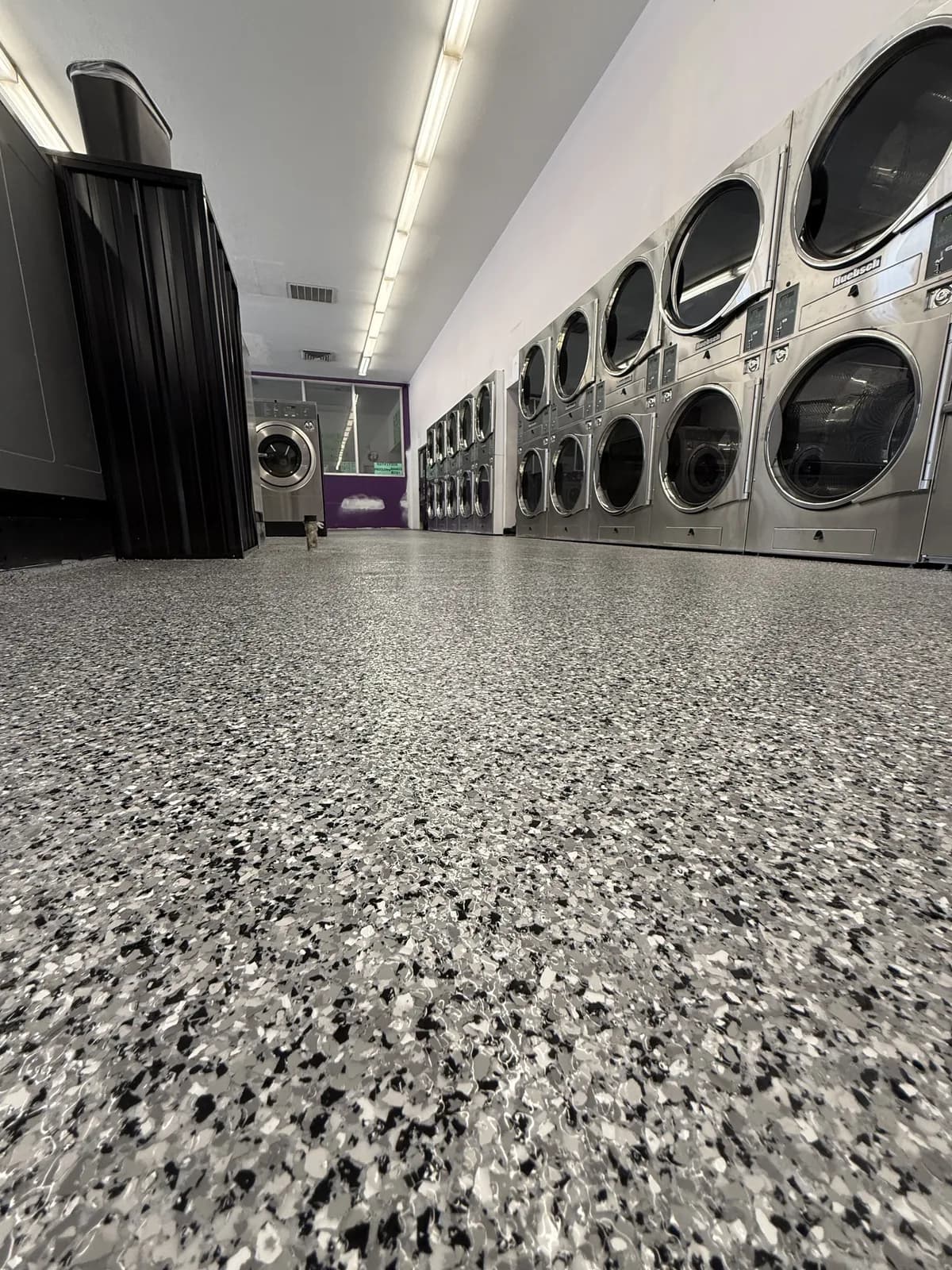 A low view along the same laundromat floor, the gray, black and white flake running the length of the room past a bank of stacked dryers