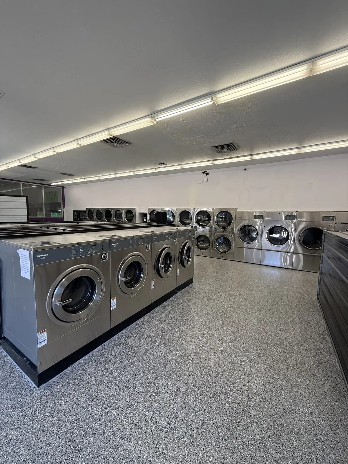 A laundromat interior with rows of stainless commercial washers and stacked dryers standing on a gray, black and white flake floor under strip lighting
