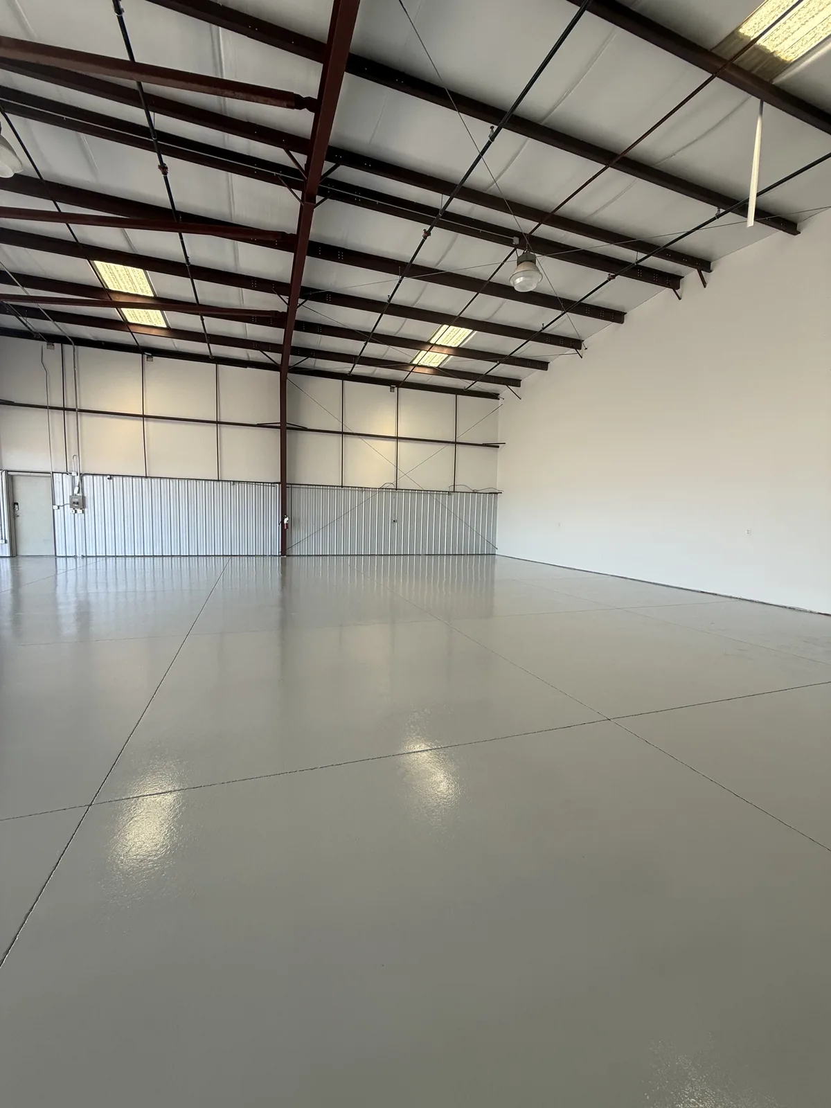 A large empty steel-framed warehouse with a plain pale gray floor coating running wall to wall, the ceiling lights reflected in its surface