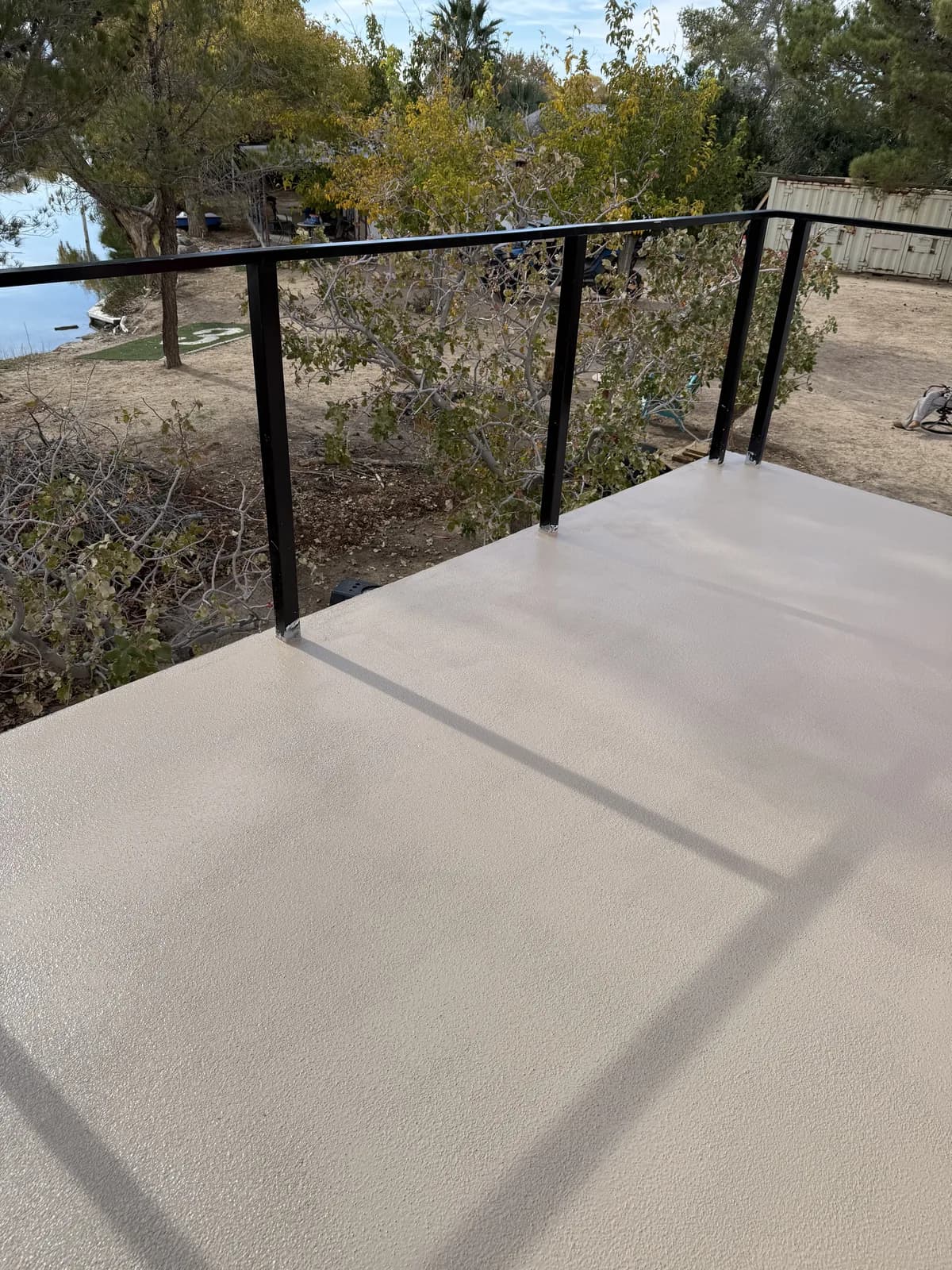 An outdoor deck finished in a pale cream coating, a black metal railing along its edge casting shadows across it, with bare trees and open water beyond
