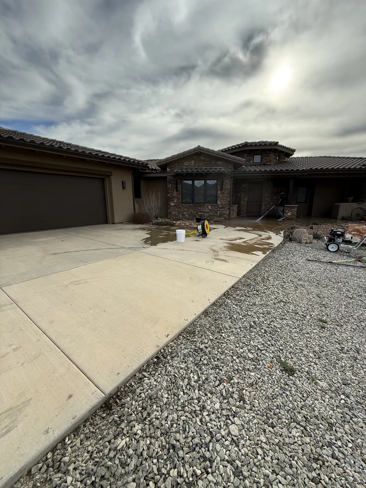 A wide residential driveway and entry being washed down, water and brown runoff spreading across the concrete, with a pressure washer and hose reel standing on the slab and a tile-roofed house behind