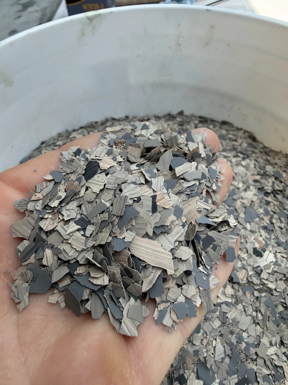 A hand holding a heap of gray, charcoal and pale tan vinyl flake chips above an open white bucket of the same