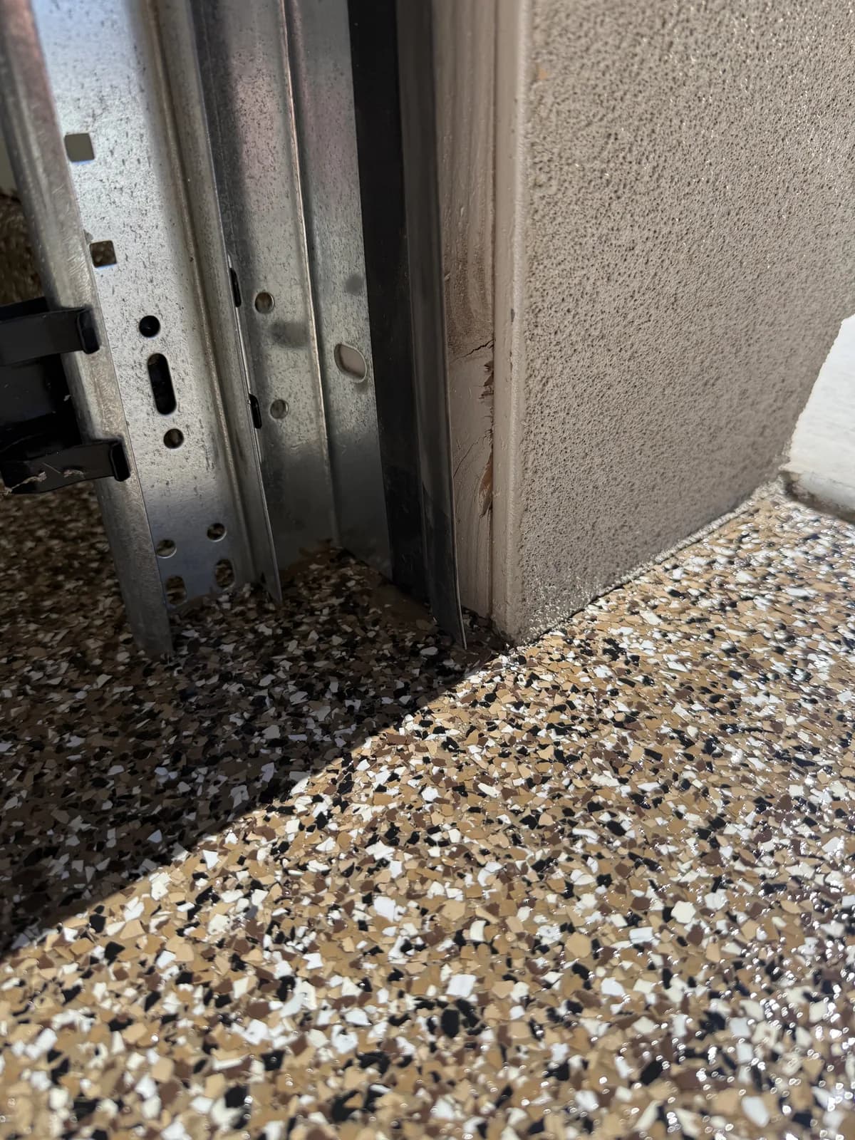 The corner where a tan flake floor meets a stucco wall and the galvanised track of a garage door, the coating running cleanly into both angles