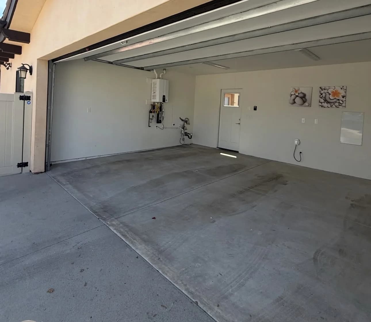 An empty garage with a bare gray concrete slab, a wall-hung tankless water heater on the back wall, a white door beside it and two small framed pictures