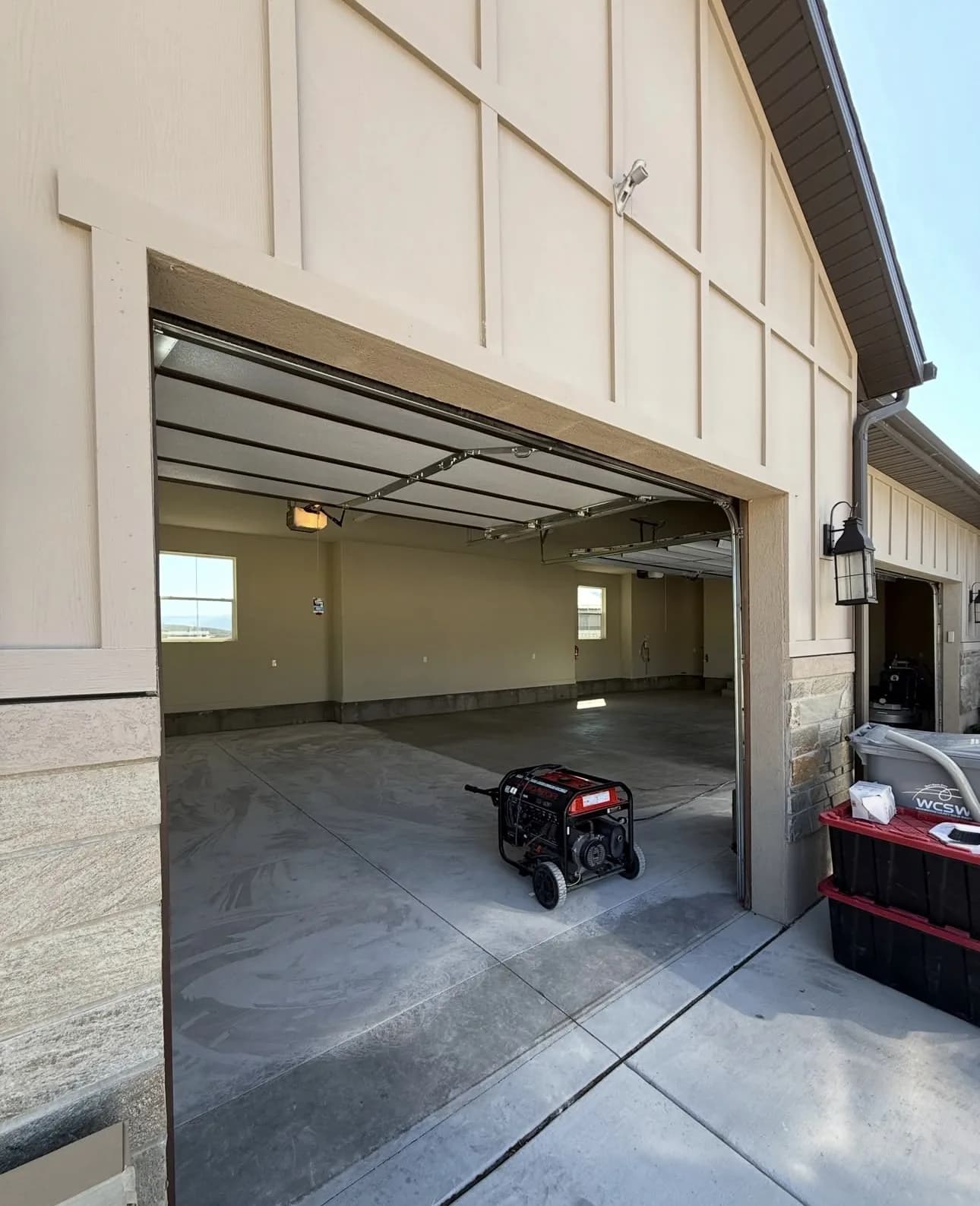 An empty garage seen from the driveway through its open door, the bare gray slab running out to meet the driveway, with a generator and a red tool chest standing just inside