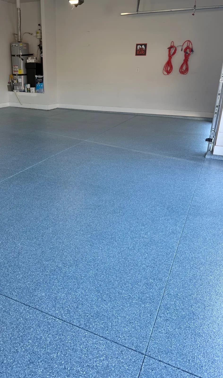 A garage floor in a blue, gray and white flake blend, with a water heater in the far corner and two red extension cords hung on the wall above