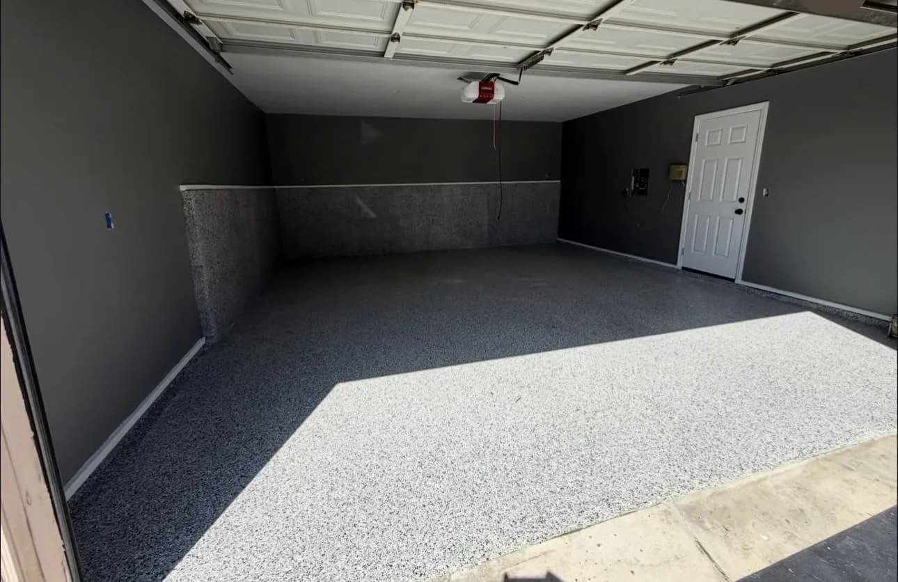 A garage with charcoal-painted walls and a gray flake floor, a bright patch of sunlight falling across it from the open door, and a white door on the right