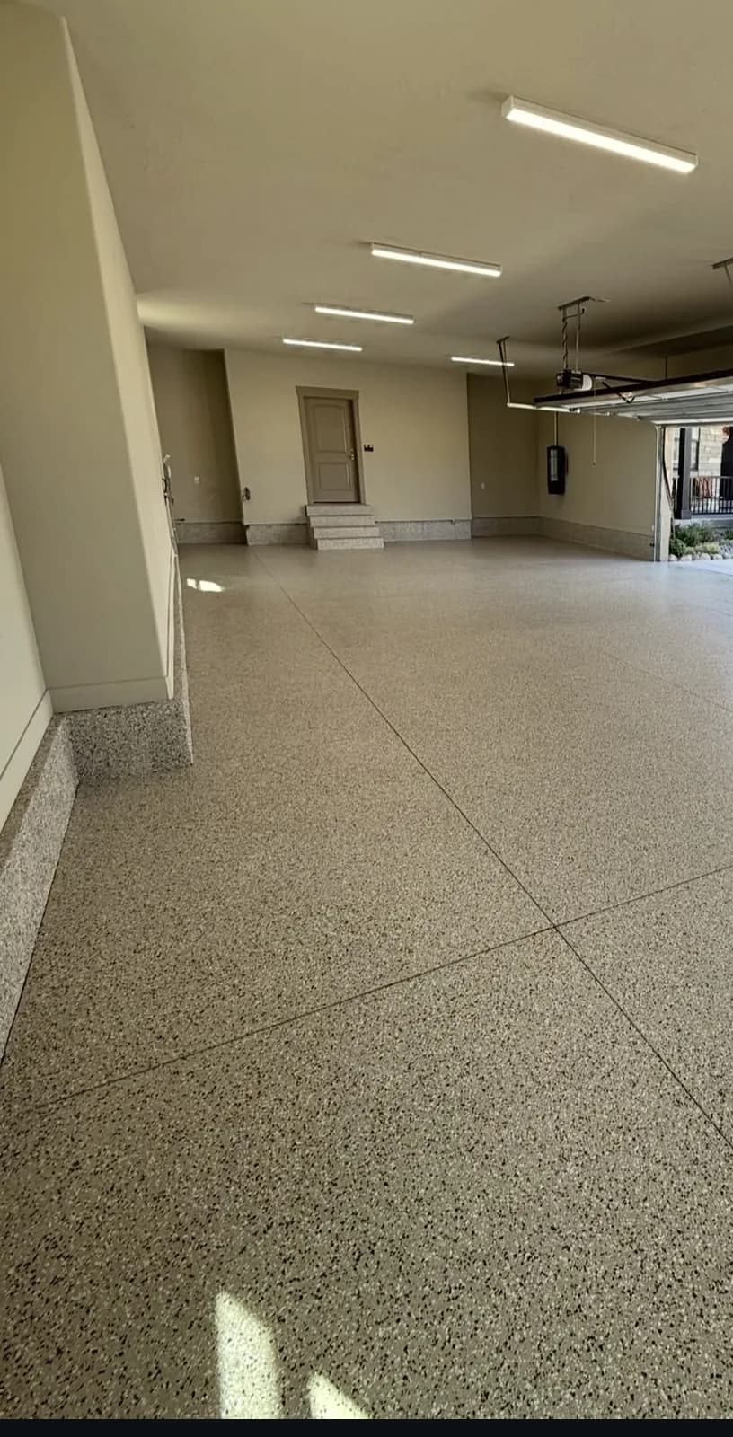 A long garage bay finished in tan flake, strip lights overhead, looking down its length to an open door and steps at the far end