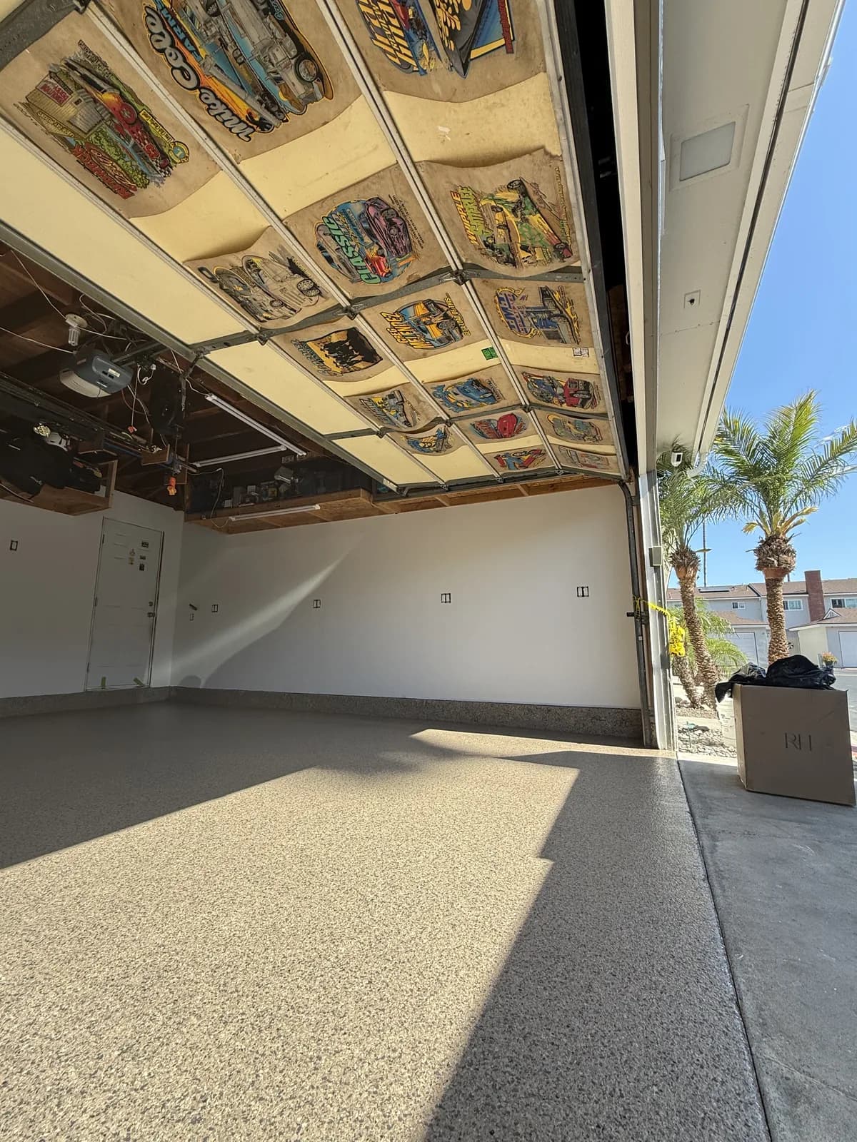 An empty garage with a pale tan flake floor and the sectional door raised, its inner face covered in framed comic and hot-rod artwork, with palm trees and a street outside