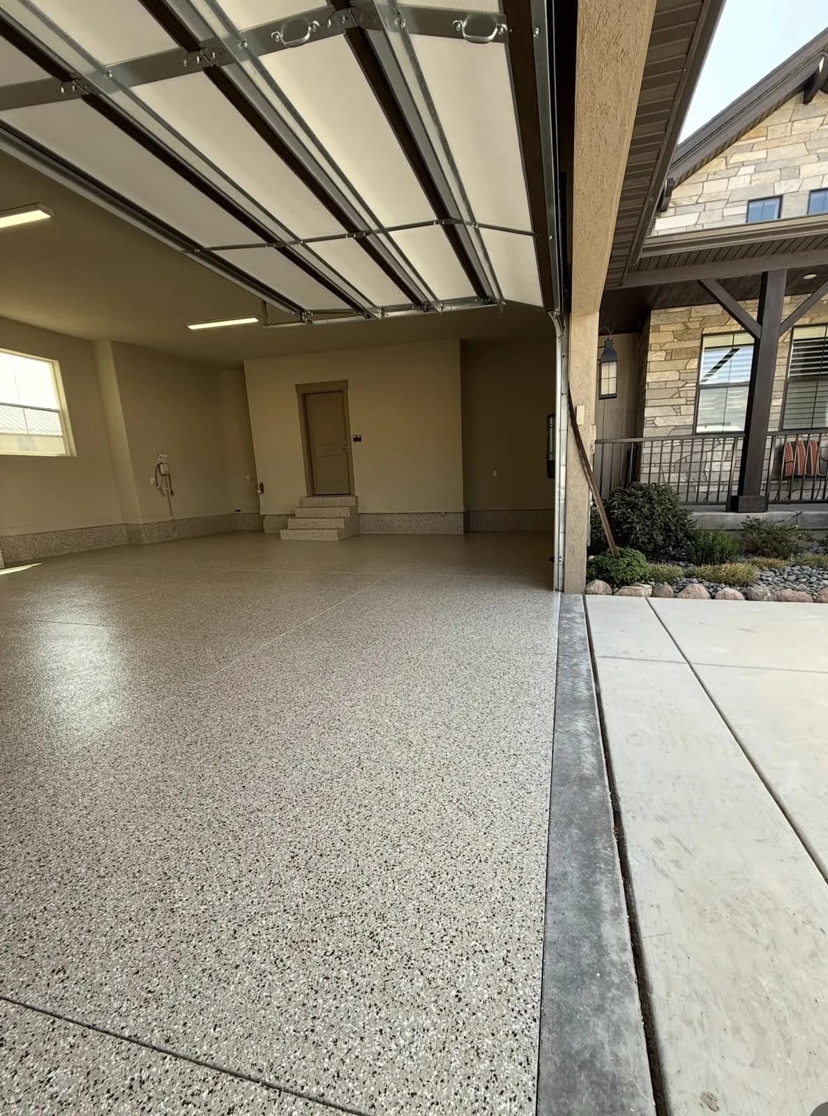 A tan flake garage floor seen from inside the bay, meeting the plain concrete driveway at the door line, with a stone-faced house across the street beyond