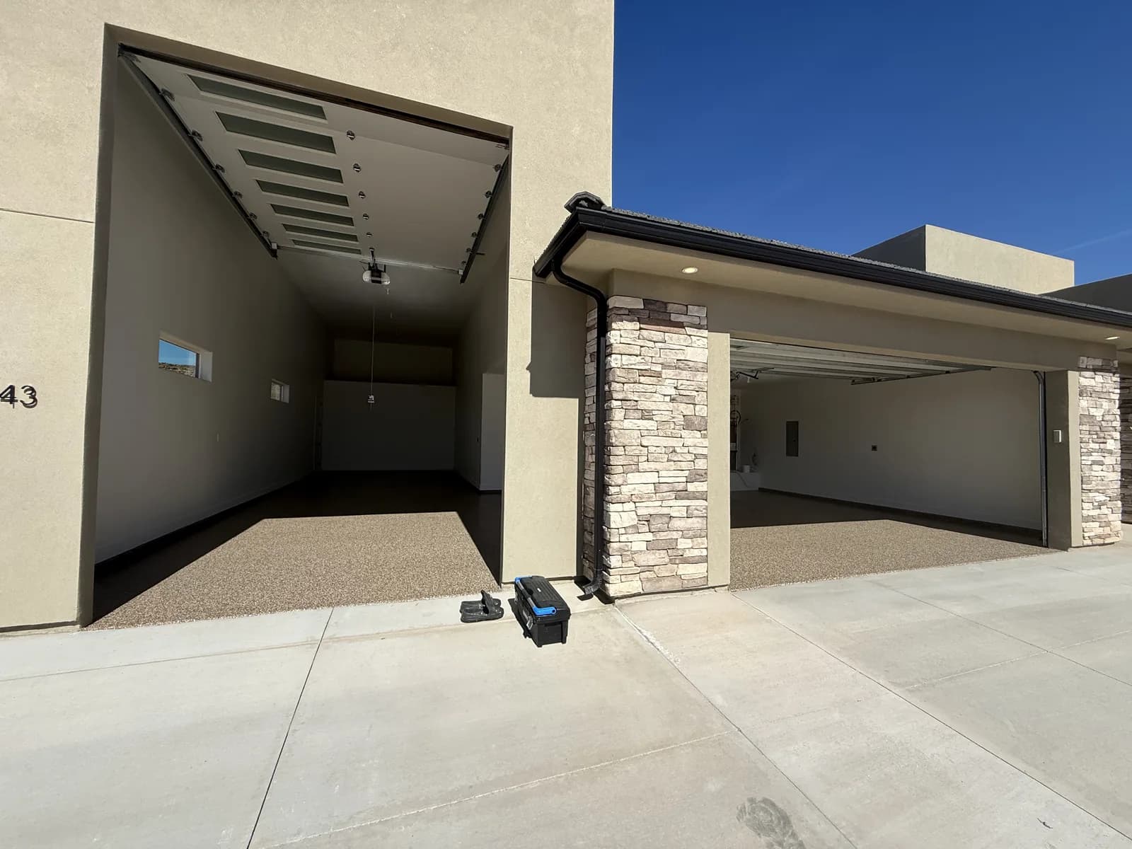 The front of a stucco and stacked-stone house with two garage doors open, showing coated flake floors inside both bays against the plain concrete driveway outside