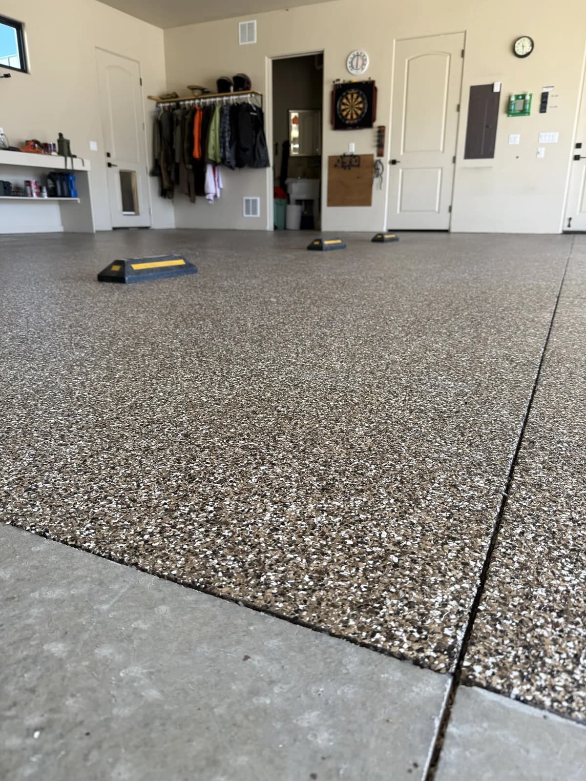 A garage in use with a gray, white and black flake floor, a rail of hanging coats and a dartboard on the far wall, a wall shelf, and exercise equipment left out on the floor