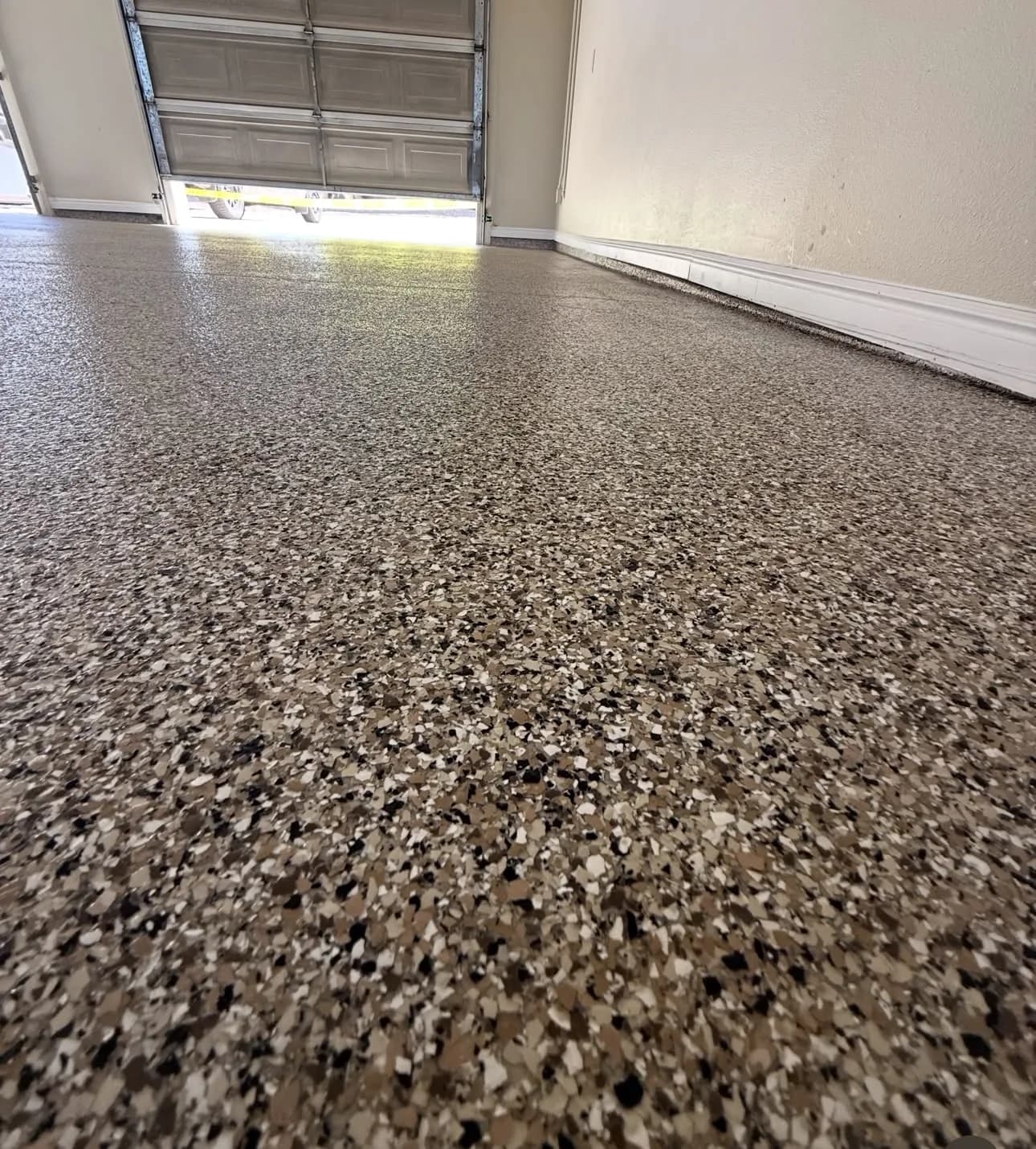 A garage floor finished in tan, brown, black and white flake, seen from floor level toward a partly open roll-up door