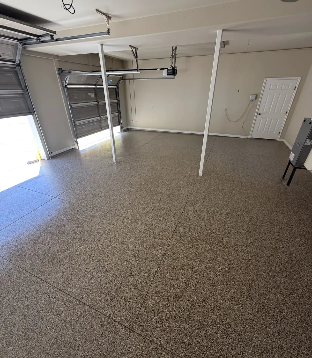 A wide garage with a brown and tan flake floor, two slim white steel posts rising from it, a sectional door raised on the left and a white door and electrical panel on the right