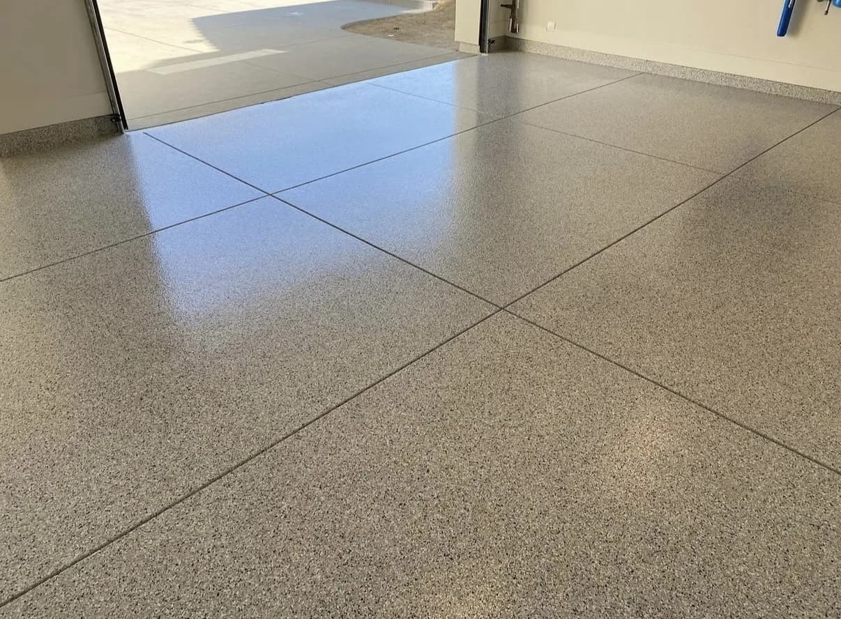 A finished garage floor in a pale gray and tan flake blend, running the depth of the bay, with the slab's saw-cut control joints still visible as fine straight lines through the coating and an open door at the far left