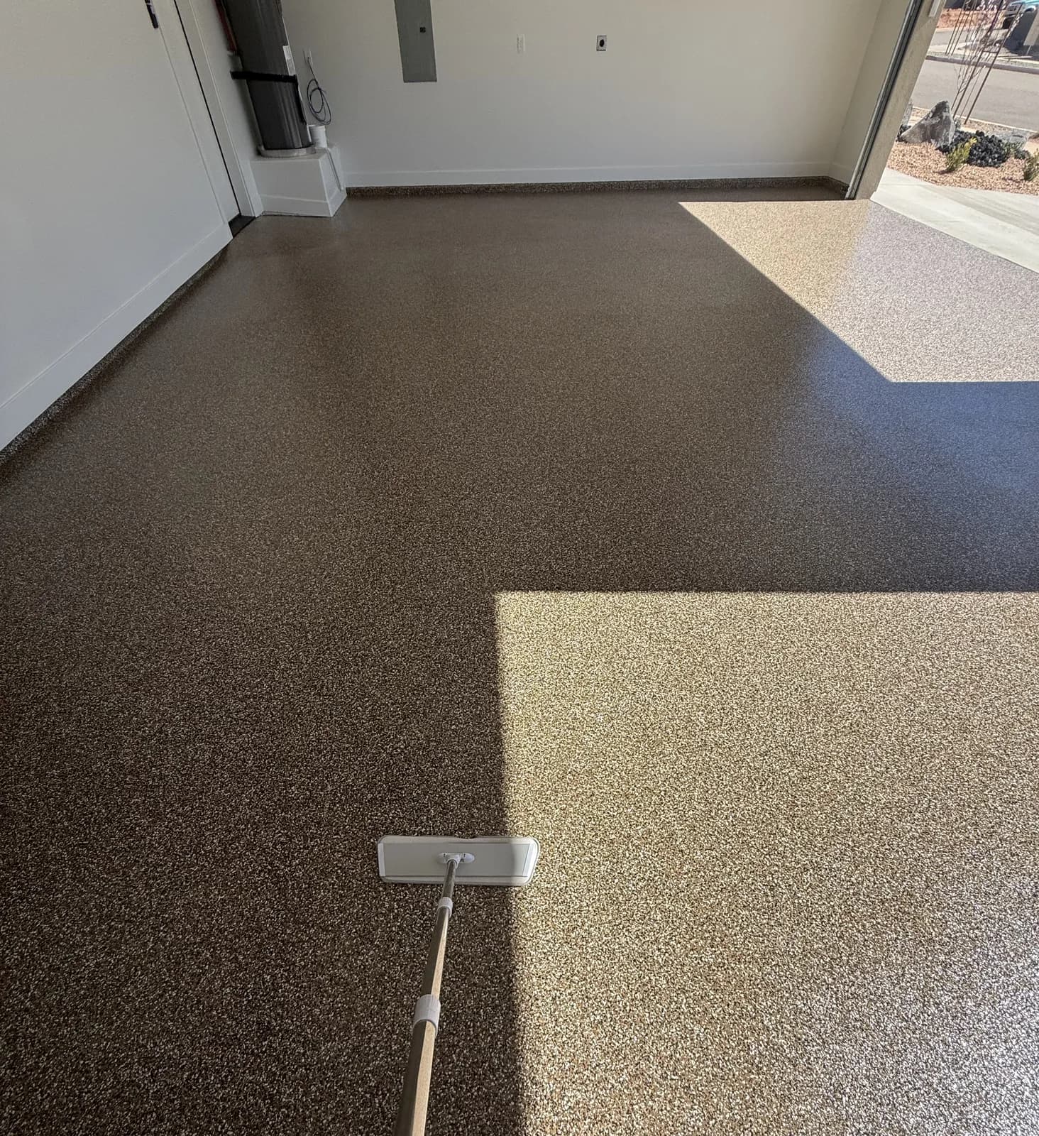 A brown flake garage floor with a hard diagonal line across it where sunlight through the open door meets shade, and a flat-head mop resting on the surface