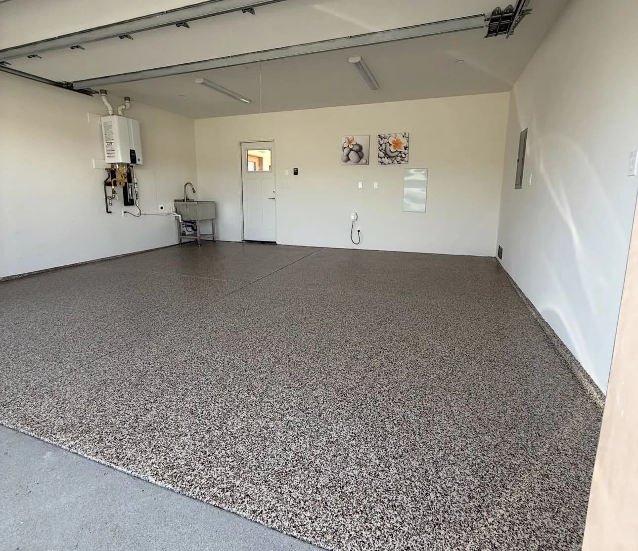A garage with a brown and tan flake floor, a wall-hung tankless water heater and utility sink on the back wall, a white door beside them and two small framed pictures
