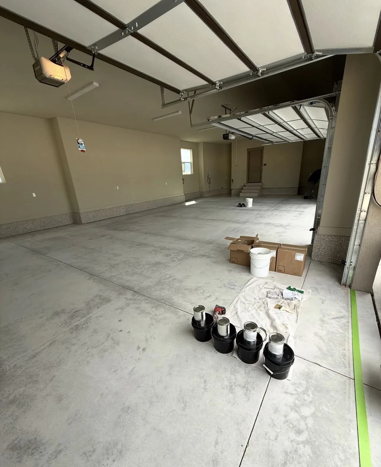 An empty garage before work, bare gray concrete with several black buckets and a dust sheet set down in the middle of the floor and a green tape line masked along the right-hand wall