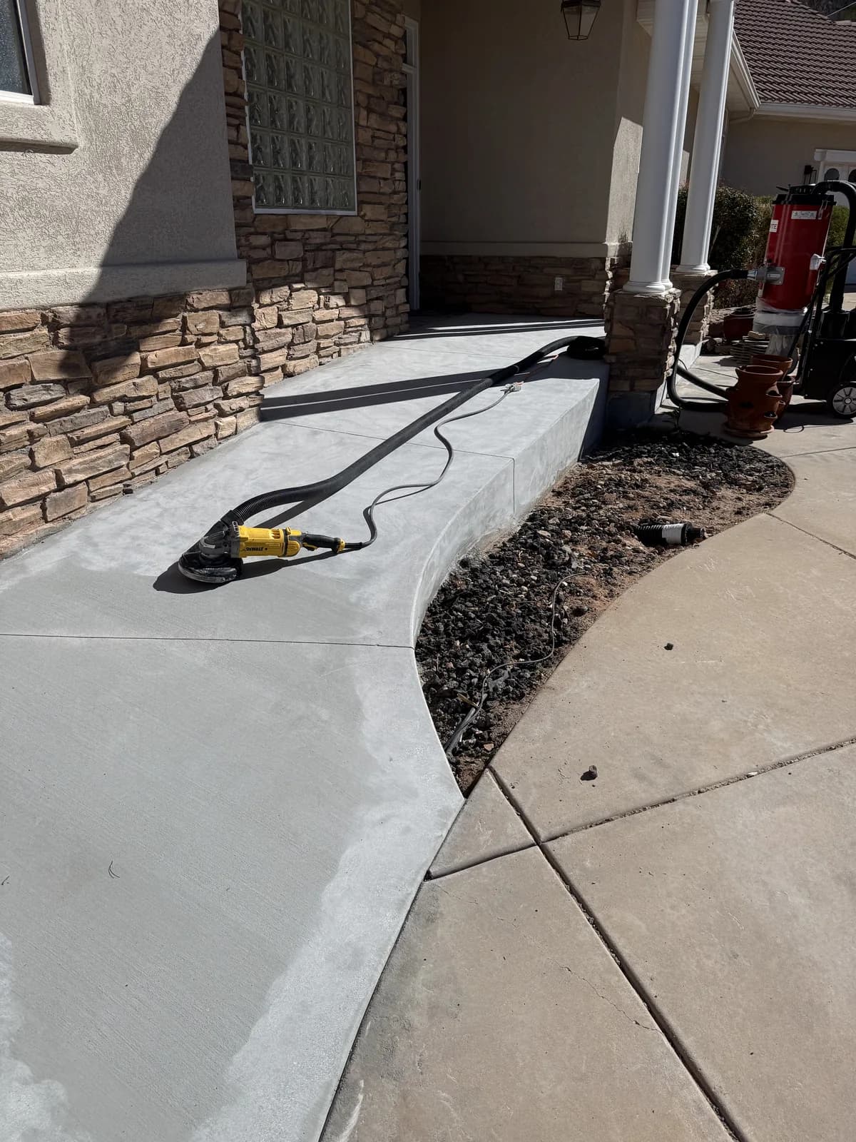A curved front walkway and step ground back to clean pale concrete, with a hand grinder and hose lying on the surface and a red dust extractor standing on the driveway beyond