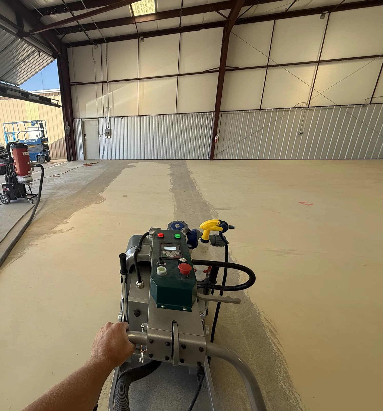 An operator's view over the handles of a walk-behind planetary grinder working a wide concrete floor inside a steel building, with a ground track visible behind the machine