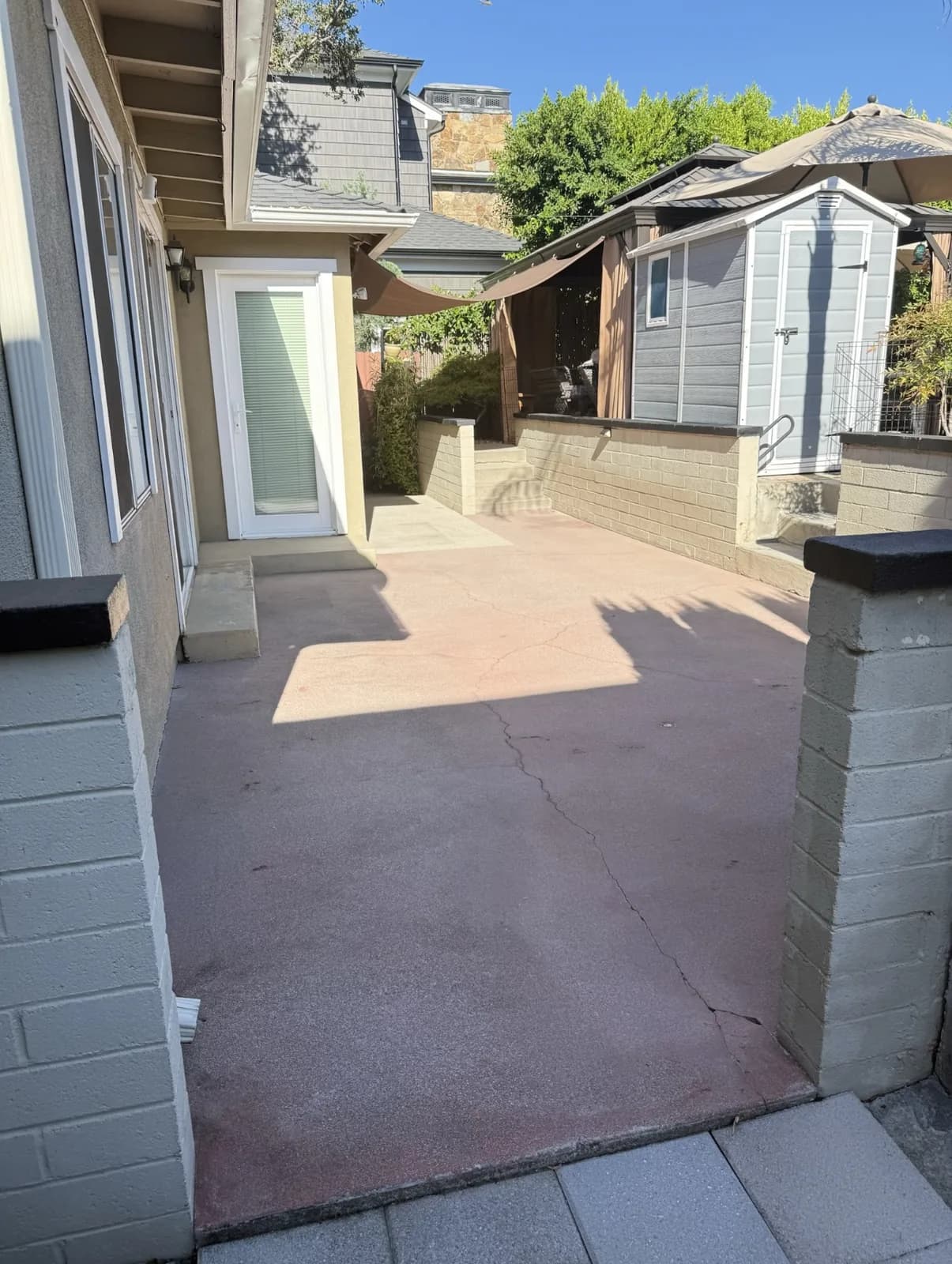 An old red-tinted concrete patio before work, faded and worn through in patches, with pale scars from earlier repairs running across it and a shed at the far end