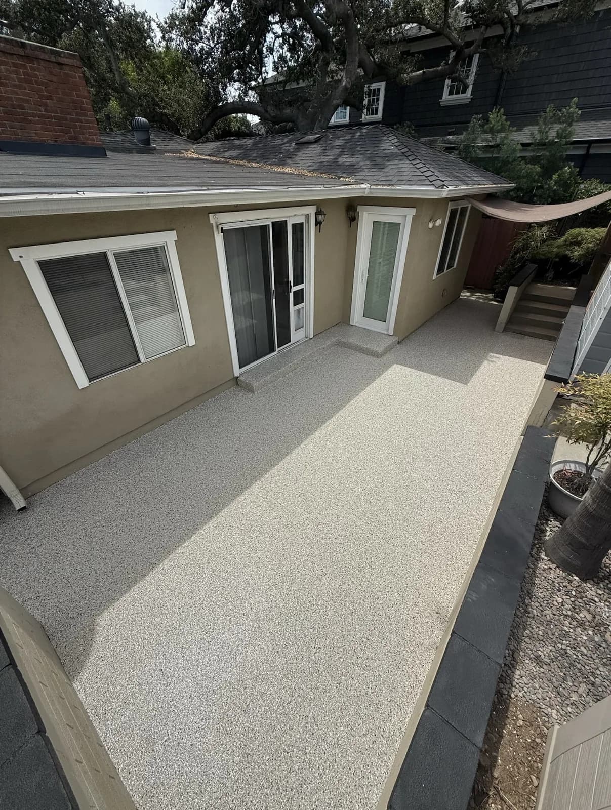 A narrow side patio finished in a pale cream and tan speckled coating, running between a stucco house wall with sliding doors and a low garden wall, with potted plants along the edge