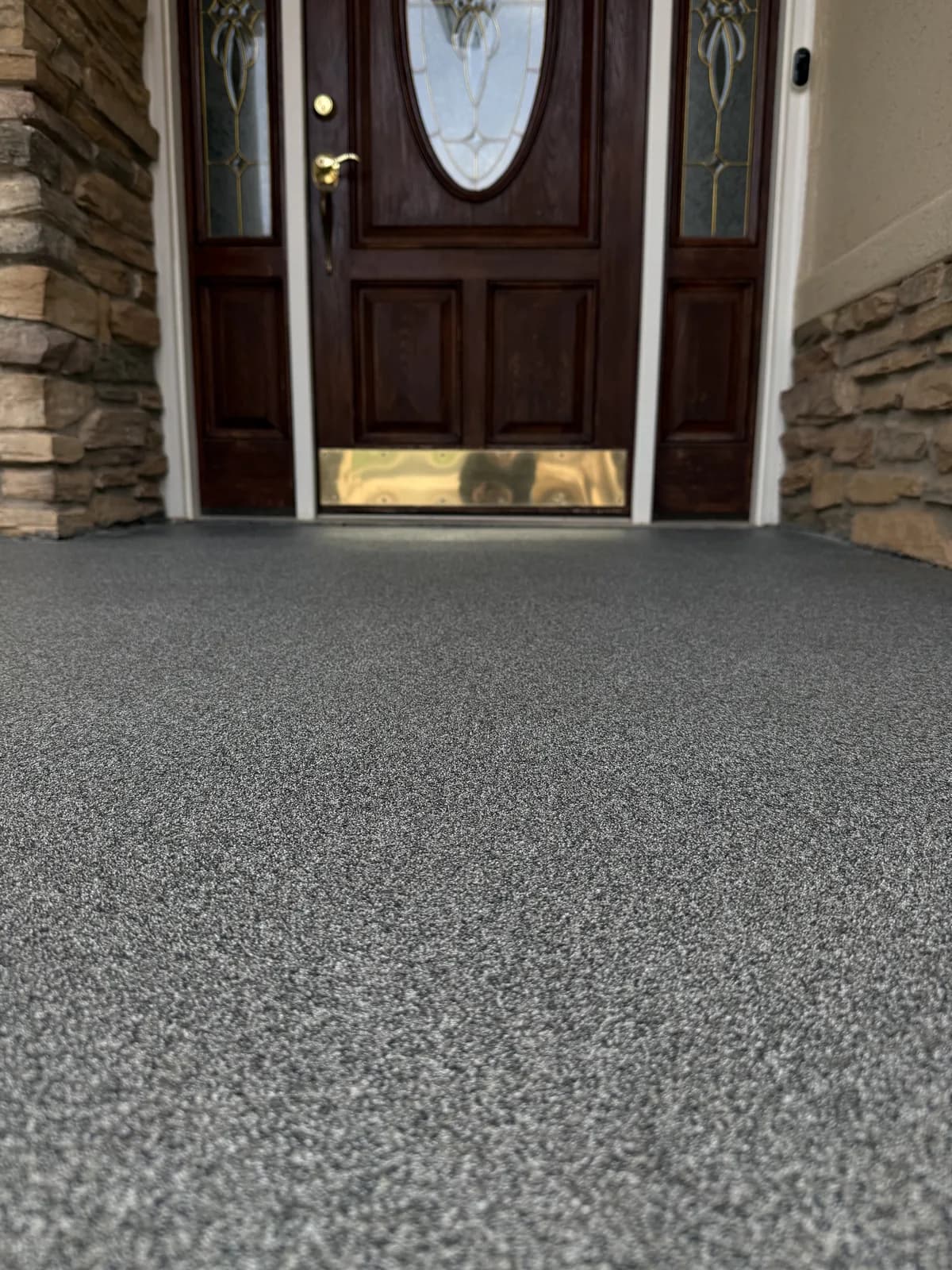 A front entry porch finished in a fine gray and white speckled coating, running up to a dark wood front door with leaded glass sidelights and stacked stone either side