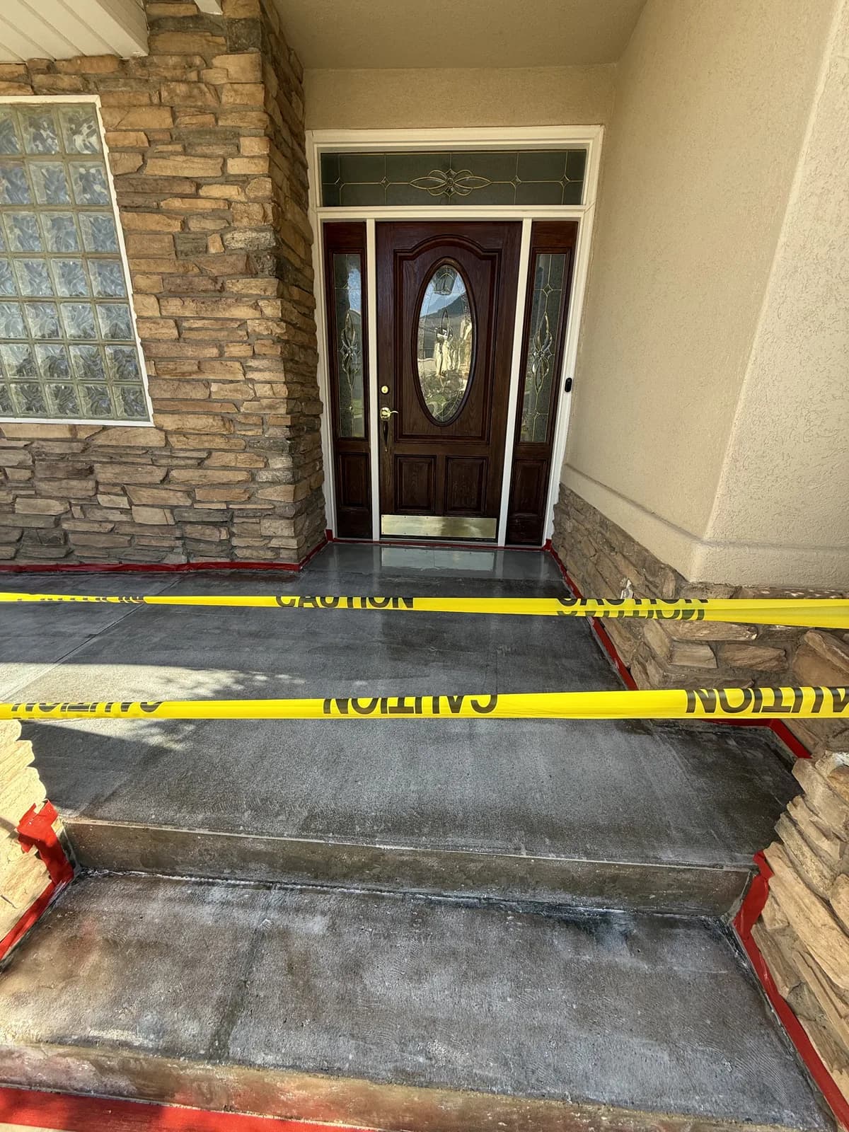 A front porch and steps prepared for coating, the stone and stucco edges masked with red tape and yellow caution tape strung across the entry, the concrete ground back to a dull gray