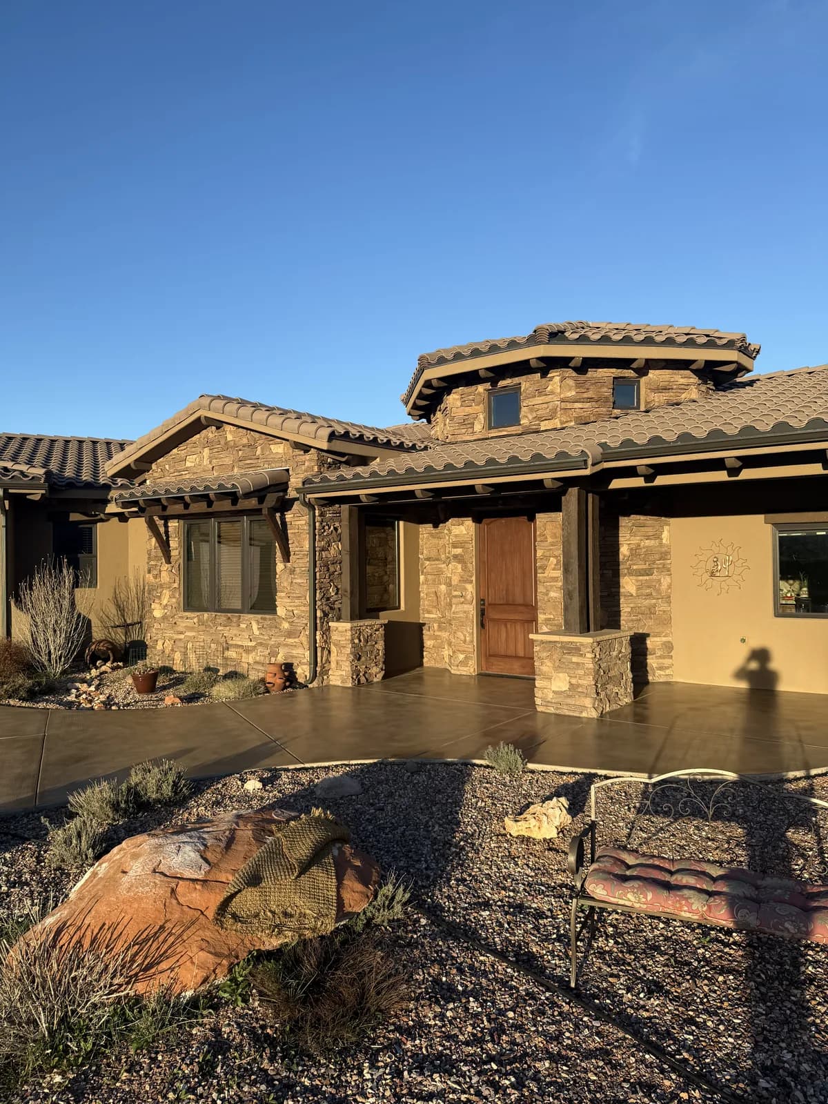 The front of a tile-roofed stucco and stone house at low sun, with a wide stained concrete entry courtyard and walkway in warm brown running to the front door
