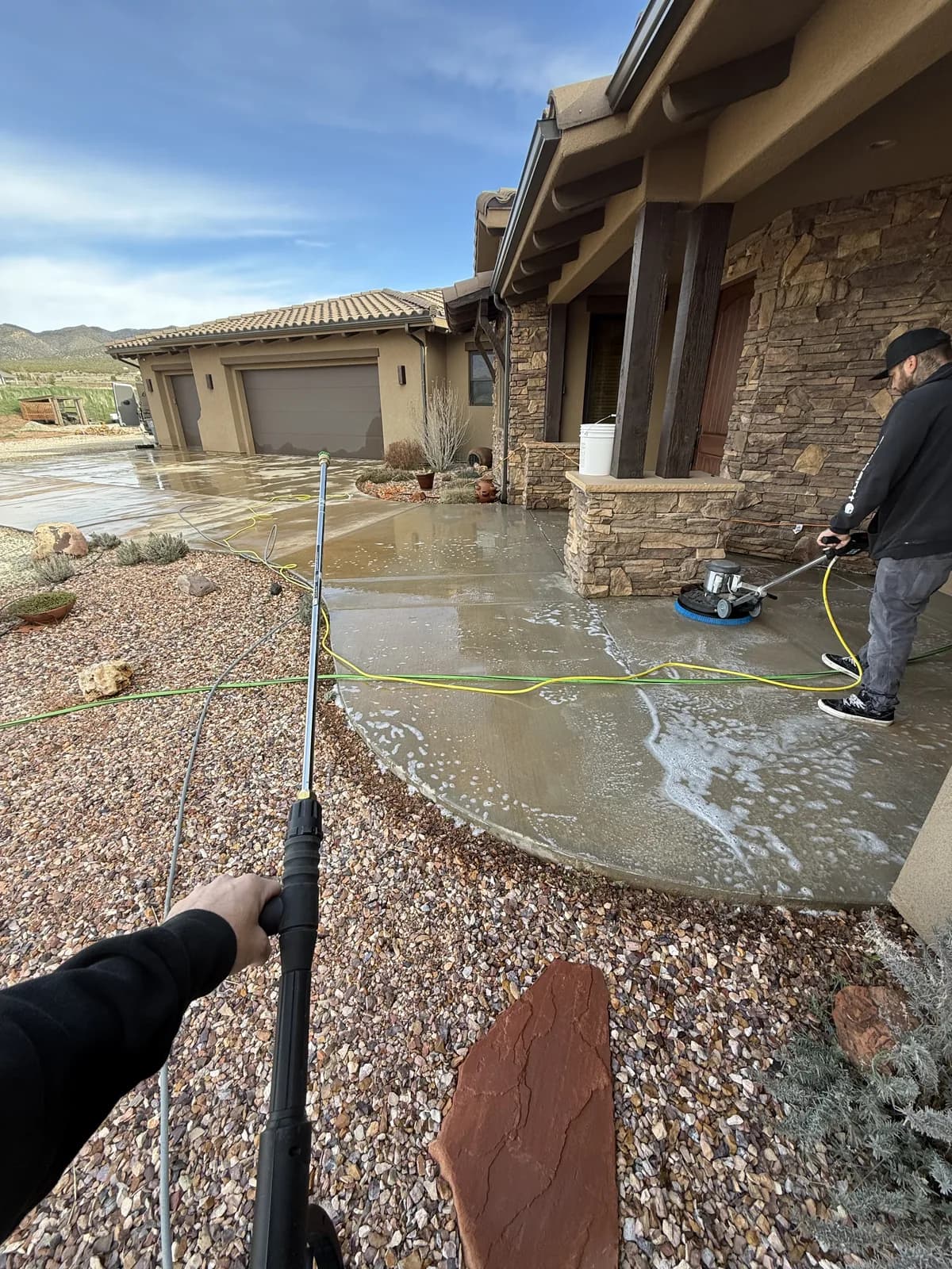 Two people cleaning a curved concrete entry with a rotary surface cleaner and a pressure wand, foam and water running across the slab, a stone-faced house and desert gravel beds around them