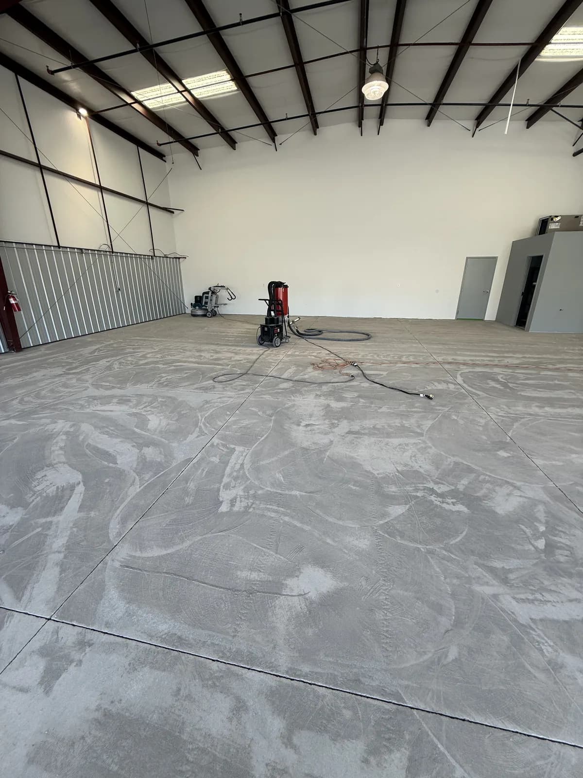 A large empty warehouse slab part-way through grinding, with wide overlapping swirl patterns across the concrete and a planetary grinder and red dust extractor parked in the middle