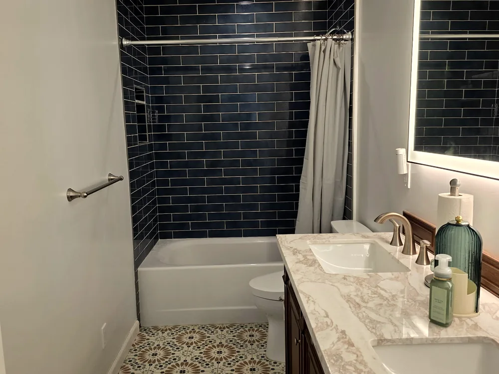 A bathroom with a dark tiled tub surround and a double marble vanity