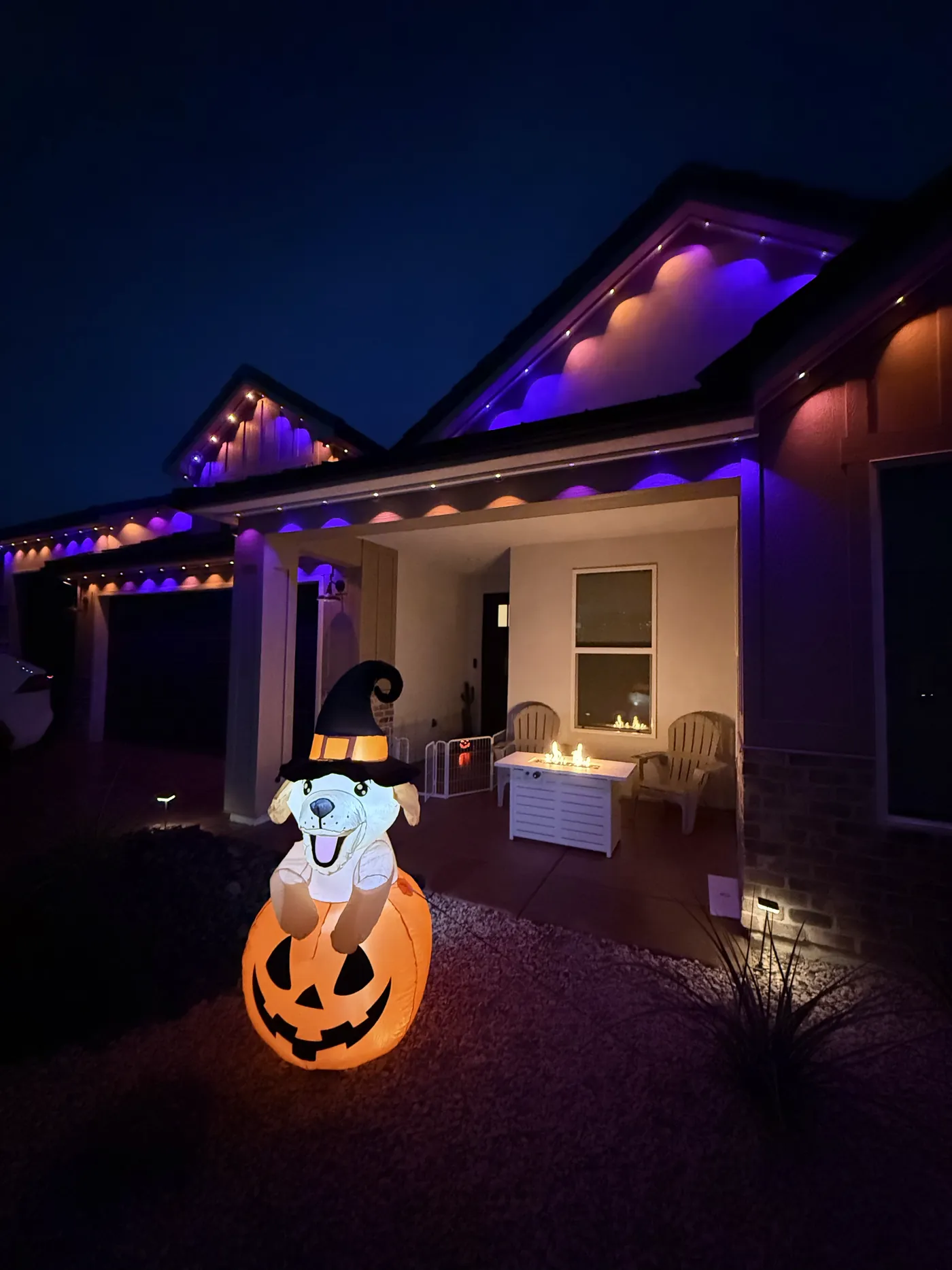 Close on the front porch after dark: small points of light along the eave and the gable, purple and orange washes on the walls, a lit fire table and two chairs.
