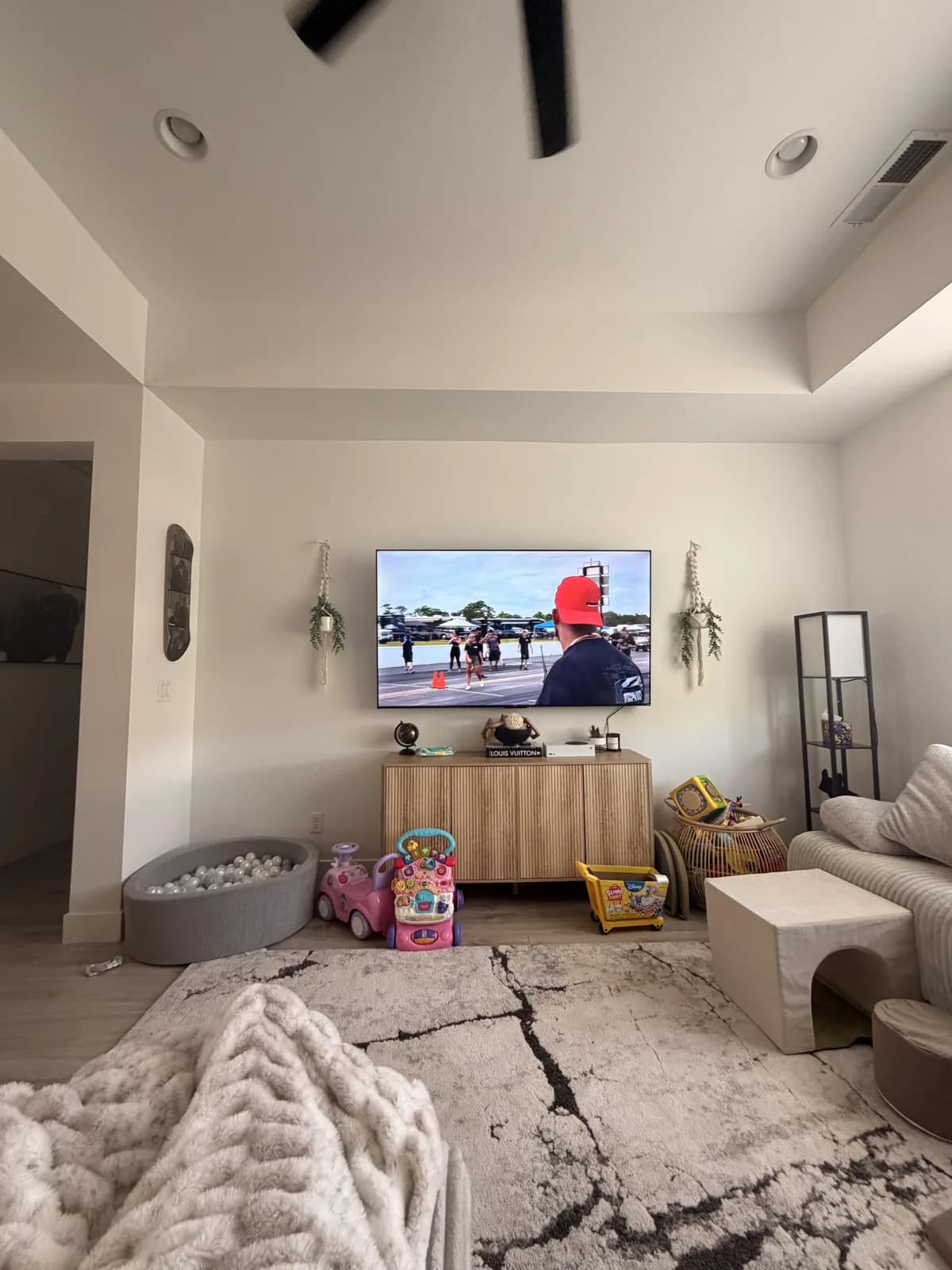 A living room with a cream-painted feature wall under a tray ceiling, a television mounted on it above a light wood console, with a rug and a ceiling fan