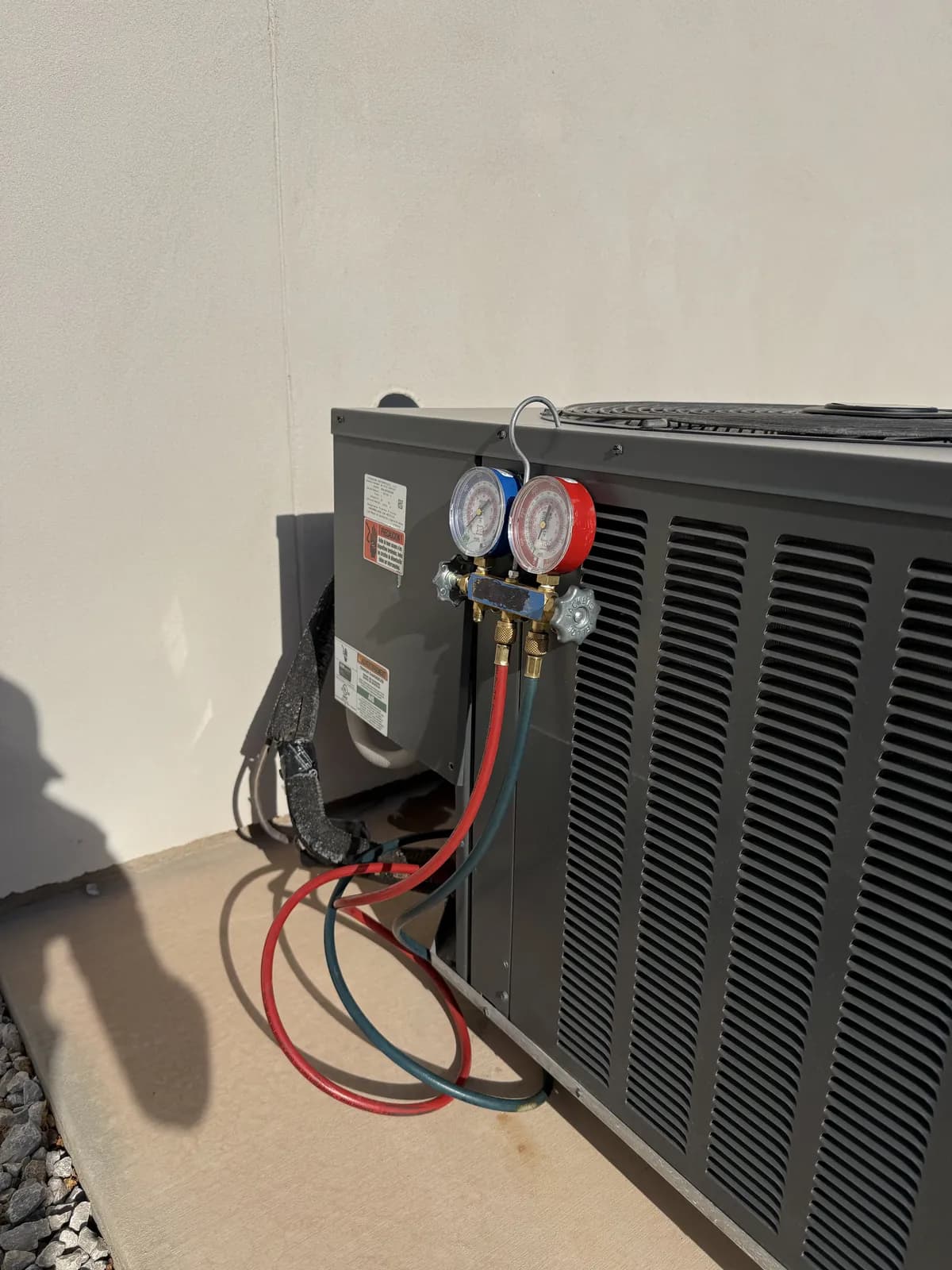 The same condenser from the side on its concrete pad against a stucco wall, the gauge set hanging from the service valves with the hoses looped below