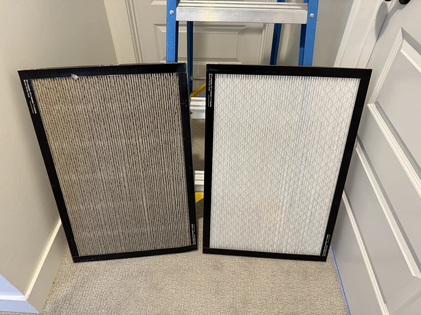 Two furnace filters standing side by side on a hallway floor: the one on the left grey and loaded with dust across its pleats, the one on the right new and white