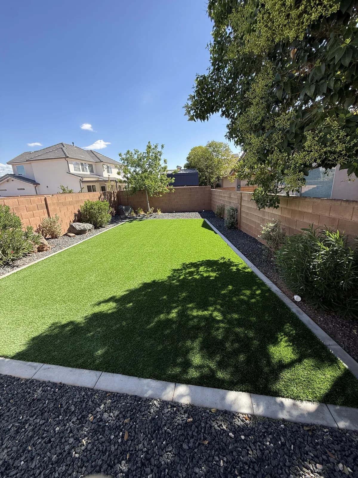 A rectangle of artificial turf edged with a concrete band in a walled back yard, gravel and shrubs along the block walls and a tree overhanging one corner