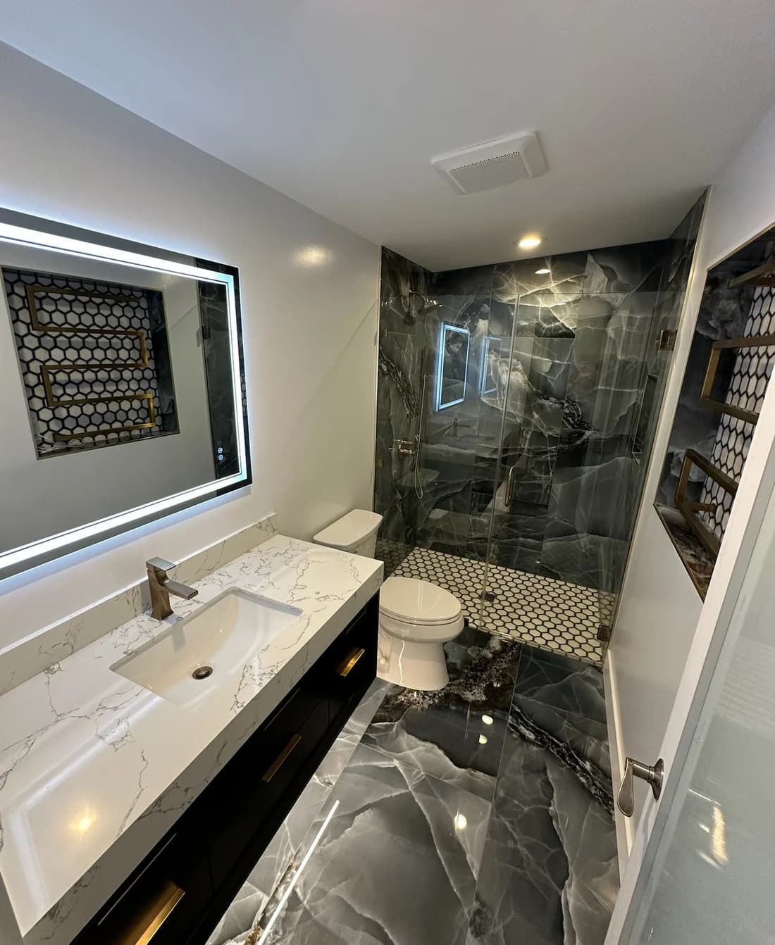 A finished bathroom: a lit mirror over a white marble vanity, and a walk-in shower in dark marble behind a glass door