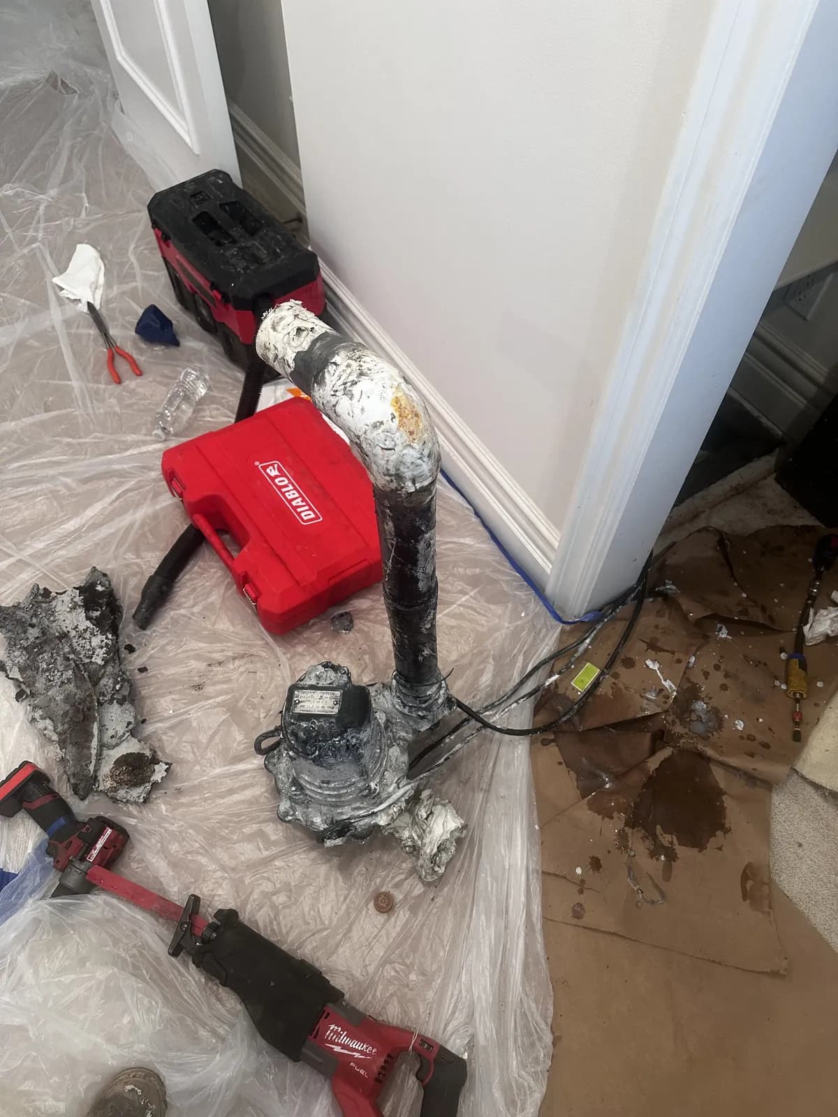 An old cast iron closet bend cut free at floor level beside the piece removed, with the floor sheeted in plastic and cutting tools laid out around it