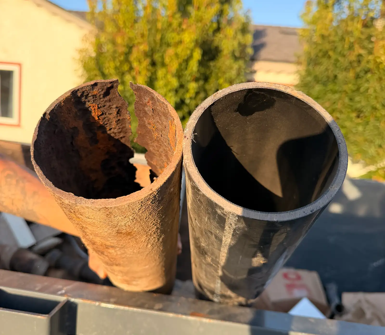A cut section of failed cast iron sewer pipe held beside a new length of the same size, the old piece split along its length with rust closing most of the bore, the new one clean and full width