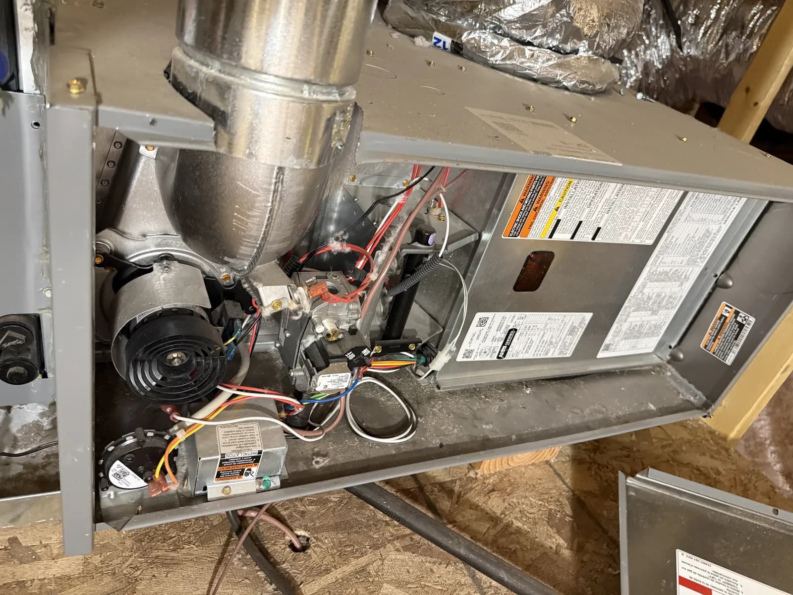 A gas furnace lying in an attic with its access panel off, showing the inducer motor, gas valve, control board and wiring, with dust across the cabinet and insulated ducts behind