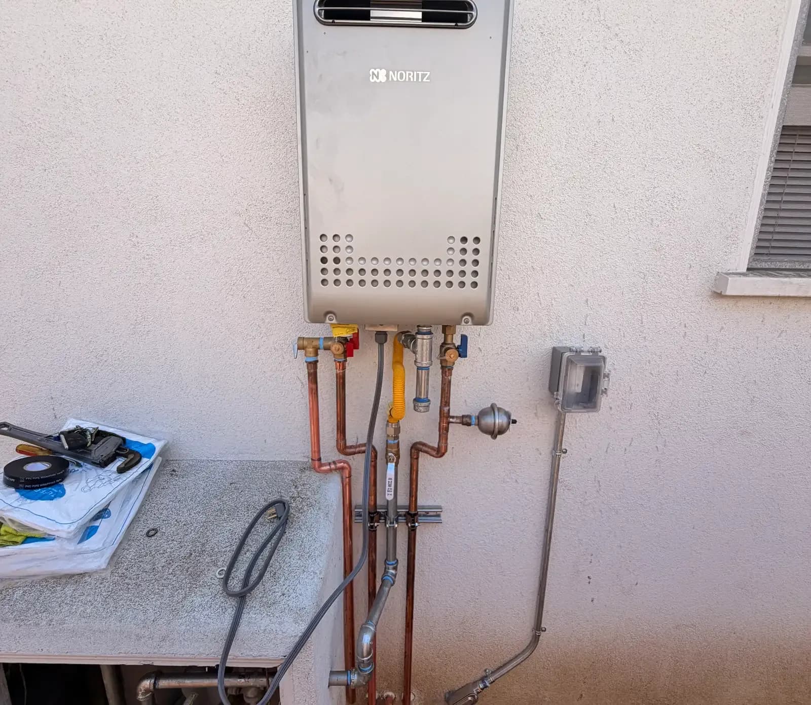 A wall-mounted tankless water heater on a stucco exterior wall with copper hot and cold lines, red and blue isolation valves, a yellow corrugated gas connector with its own shutoff, a condensate drain and a weatherproof electrical disconnect