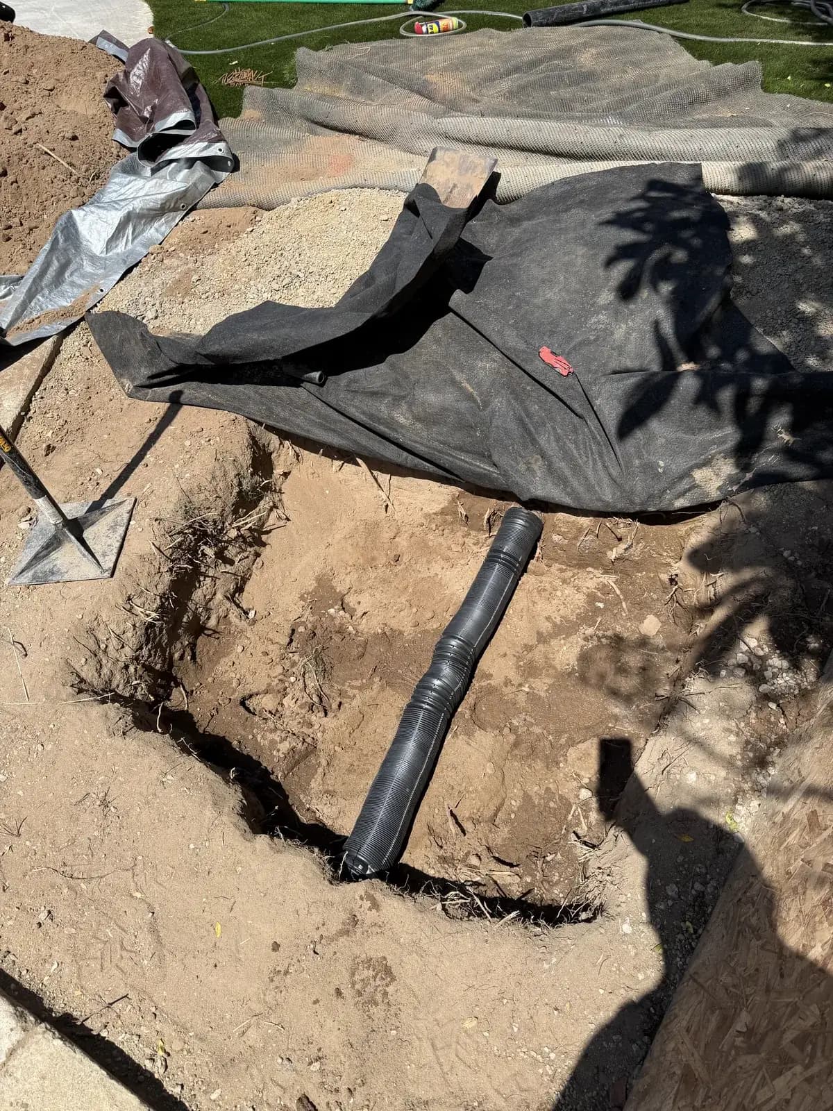 An excavated pit with new pipe laid alongside it and protective sheeting over the surrounding ground