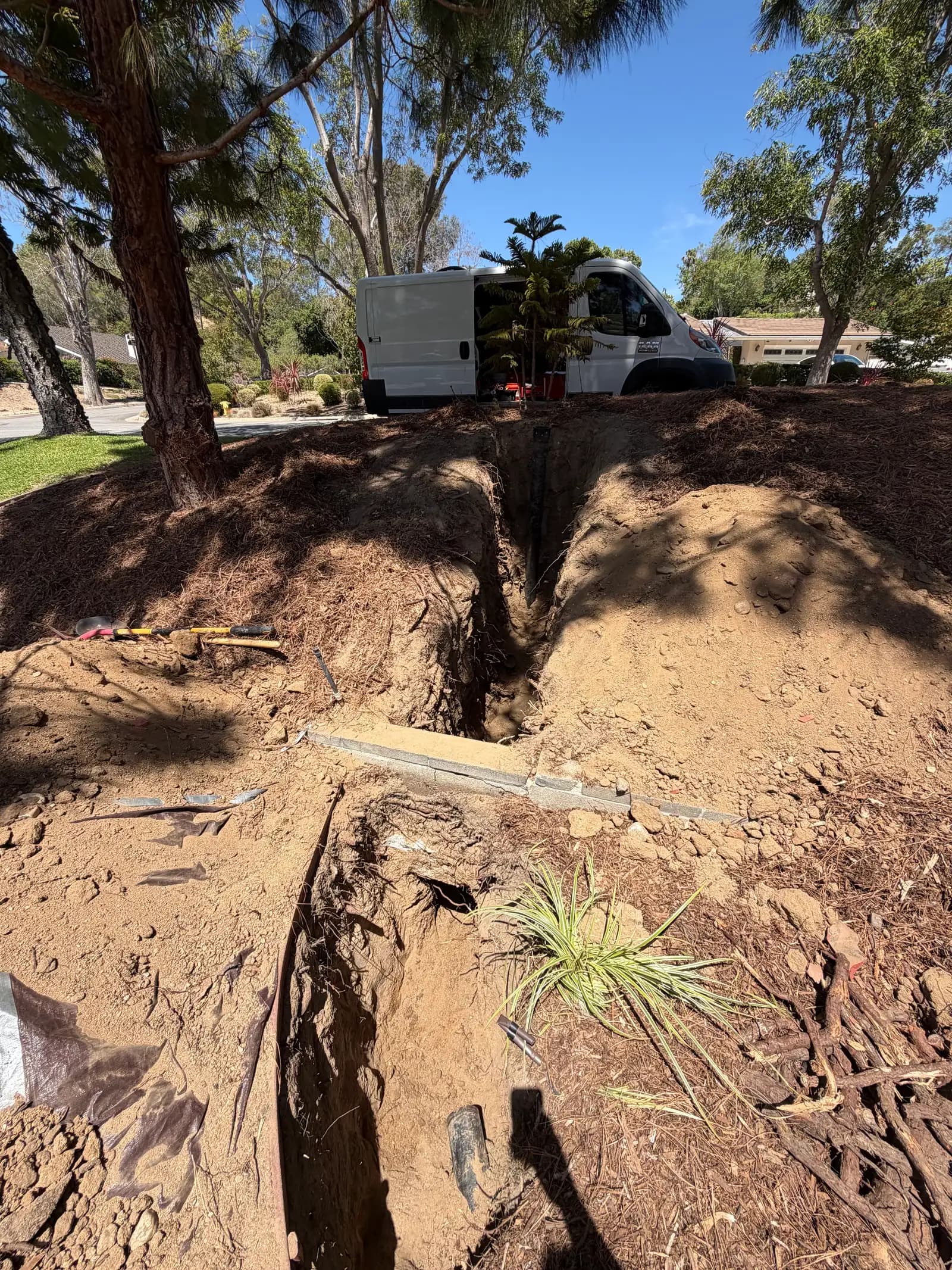 An open trench for a sewer lateral cutting across a landscaped front yard, spoil piled either side and tree roots cut through