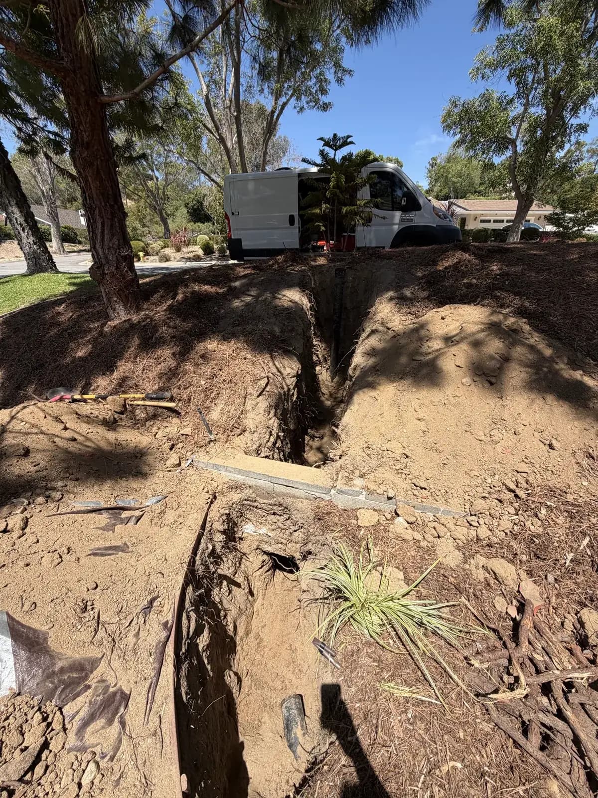 An open trench cut across a yard for a sewer replacement, with spoil piled beside it